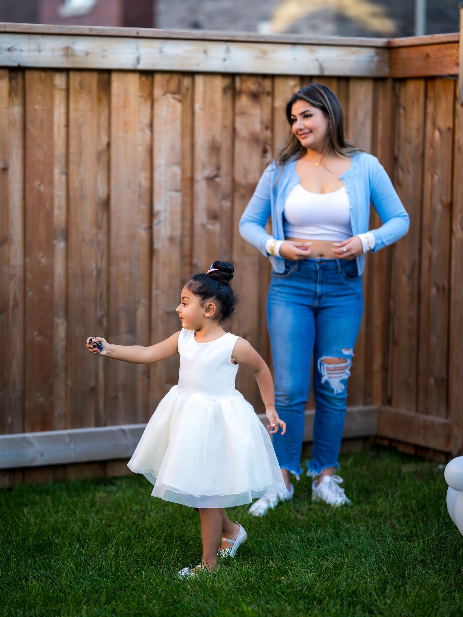 A woman in blue cardigan and jeans watches a little girl in white dress twirl in a backyard.
