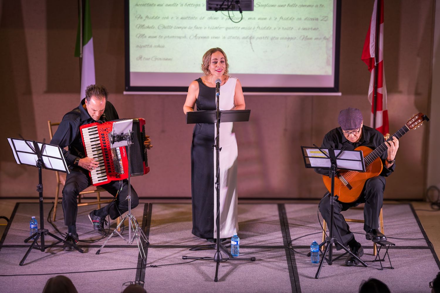 A female vocalist performs with an accordion player and guitarist on stage at a formal musical event.