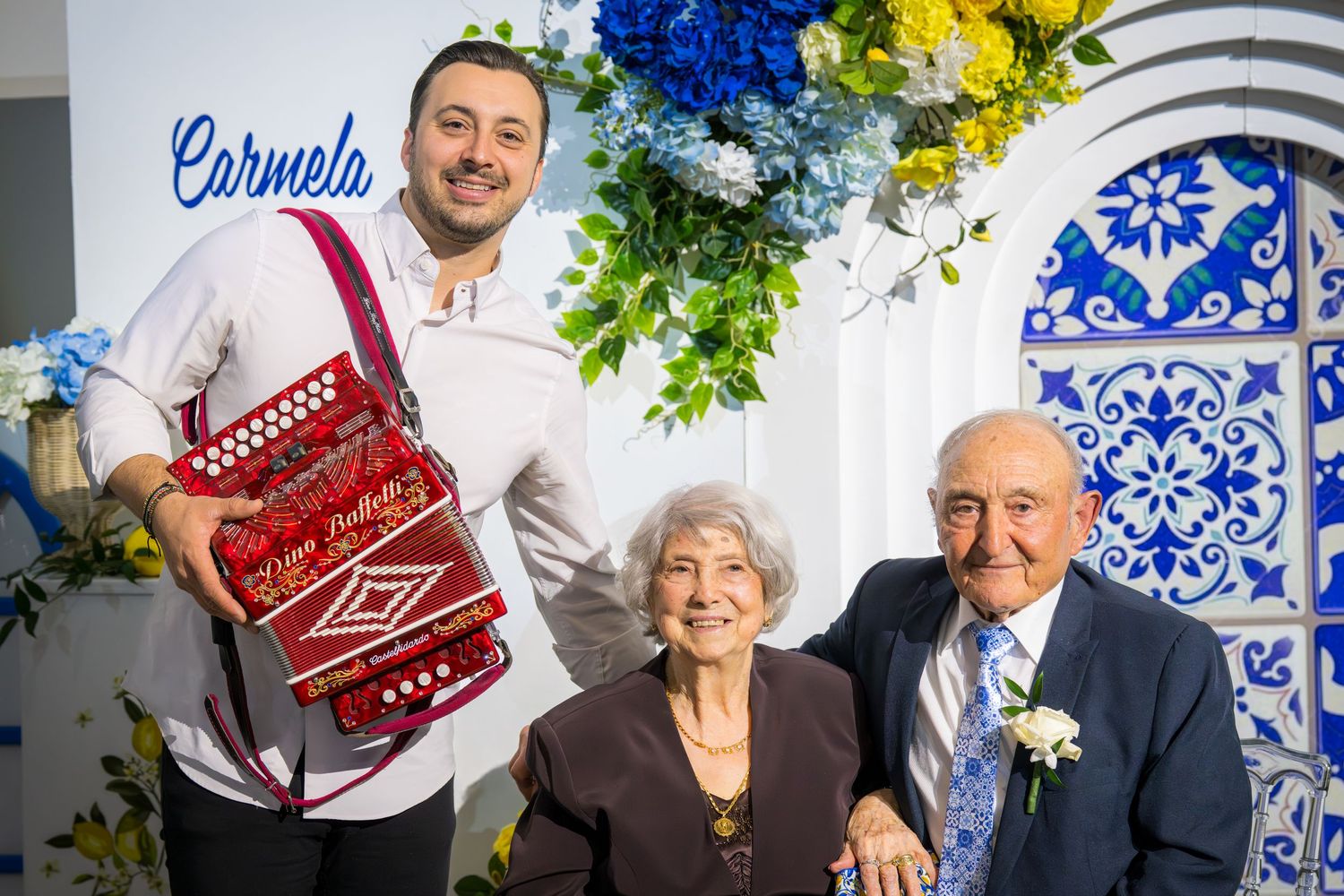 Elderly couple poses with accordion player at Carmela anniversary celebration with blue floral backdrop.