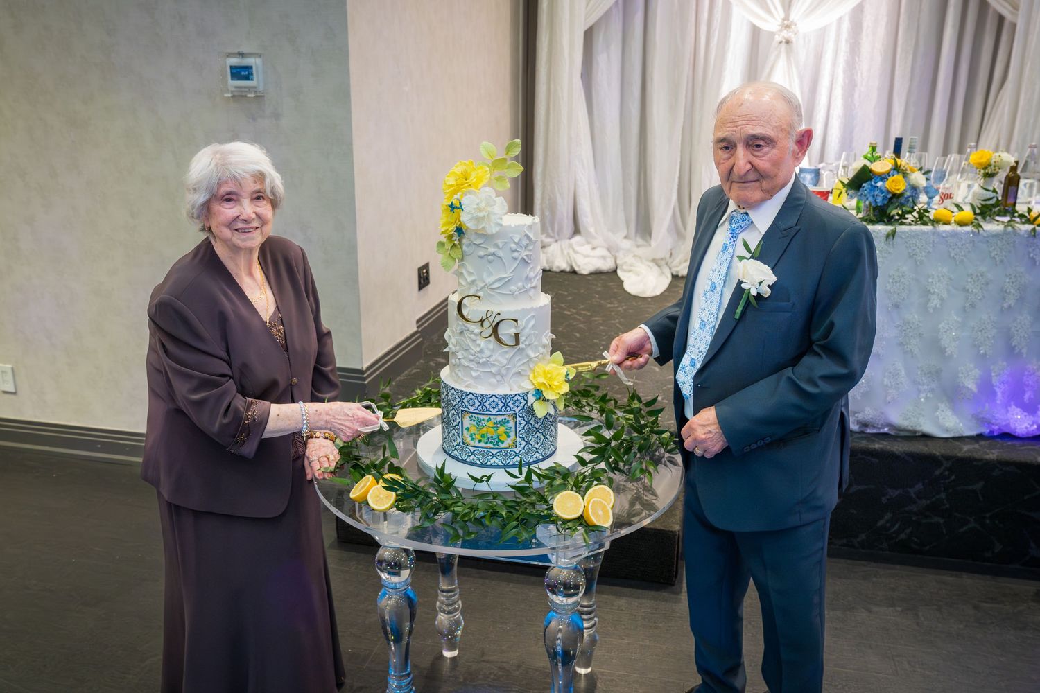 Elderly couple cutting tiered wedding anniversary cake decorated with lemons and monogram.