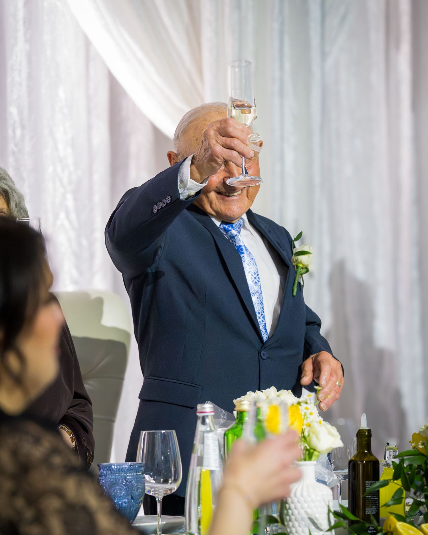 Elderly man in navy suit raising champagne glass for a toast at a wedding reception.