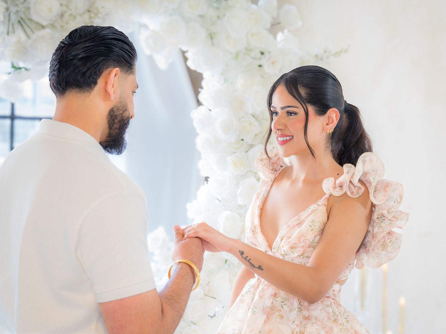 Couple holding hands and smiling in front of a white floral arch during proposal celebration.
