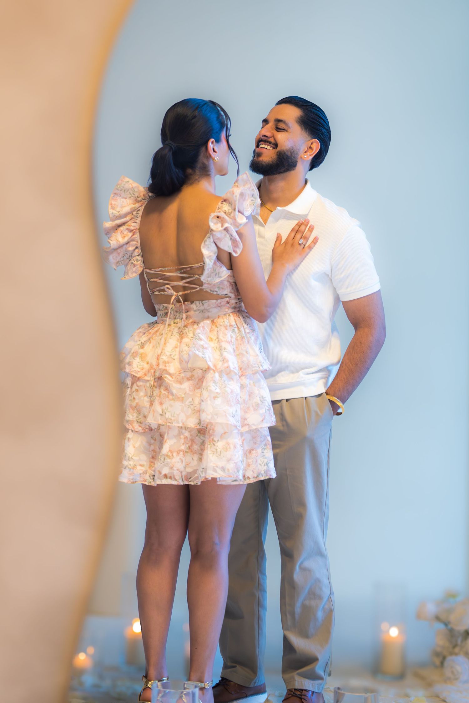 Smiling couple embracing in romantic candle-lit proposal setting, woman wearing backless floral mini dress.