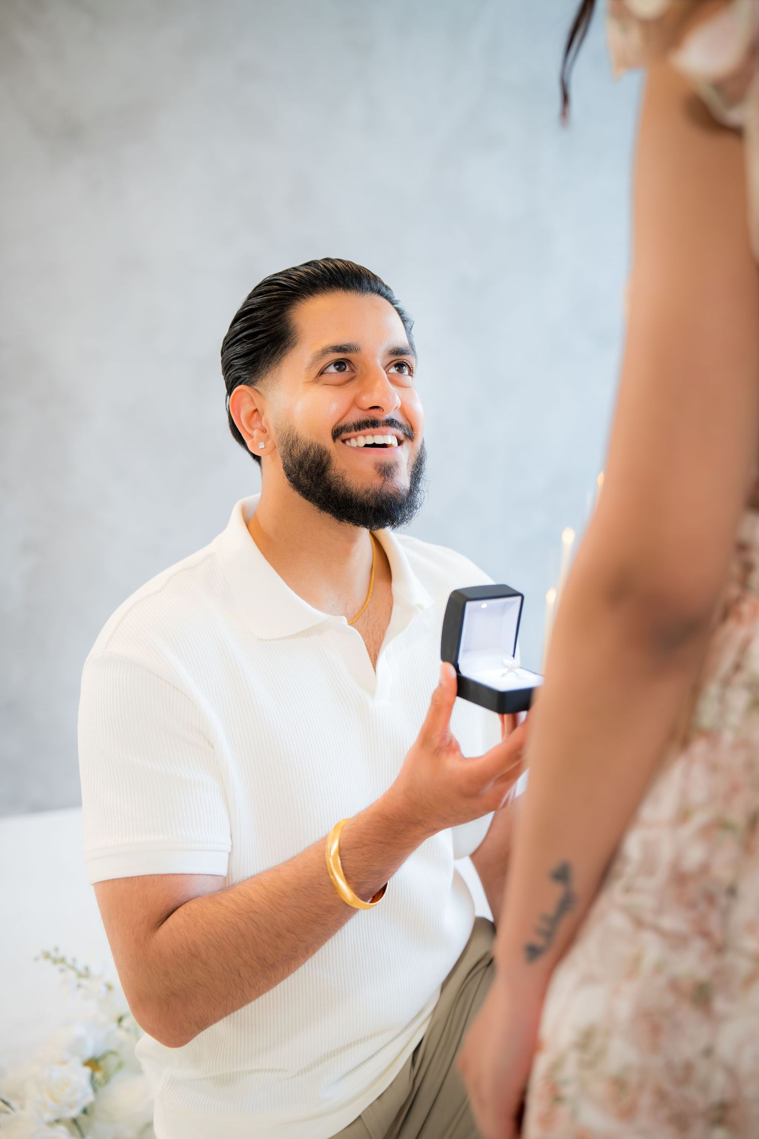 Man on one knee smiling while presenting engagement ring box to his partner.
