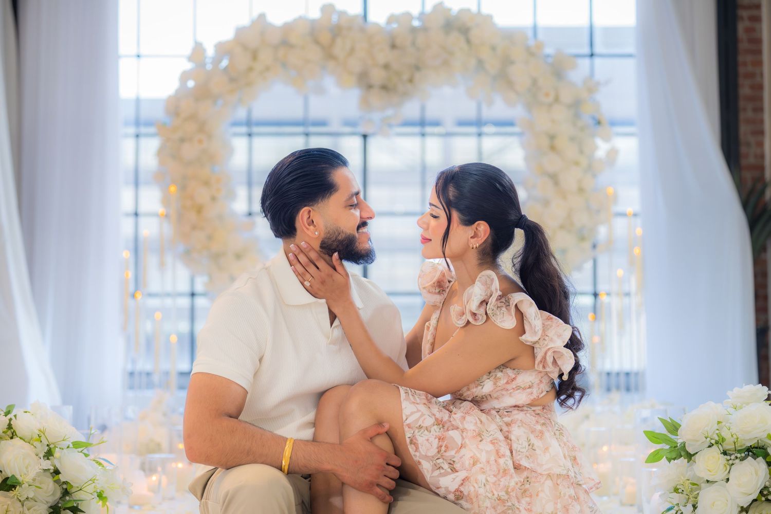 Romantic couple sitting together beneath a white floral heart-shaped arch.