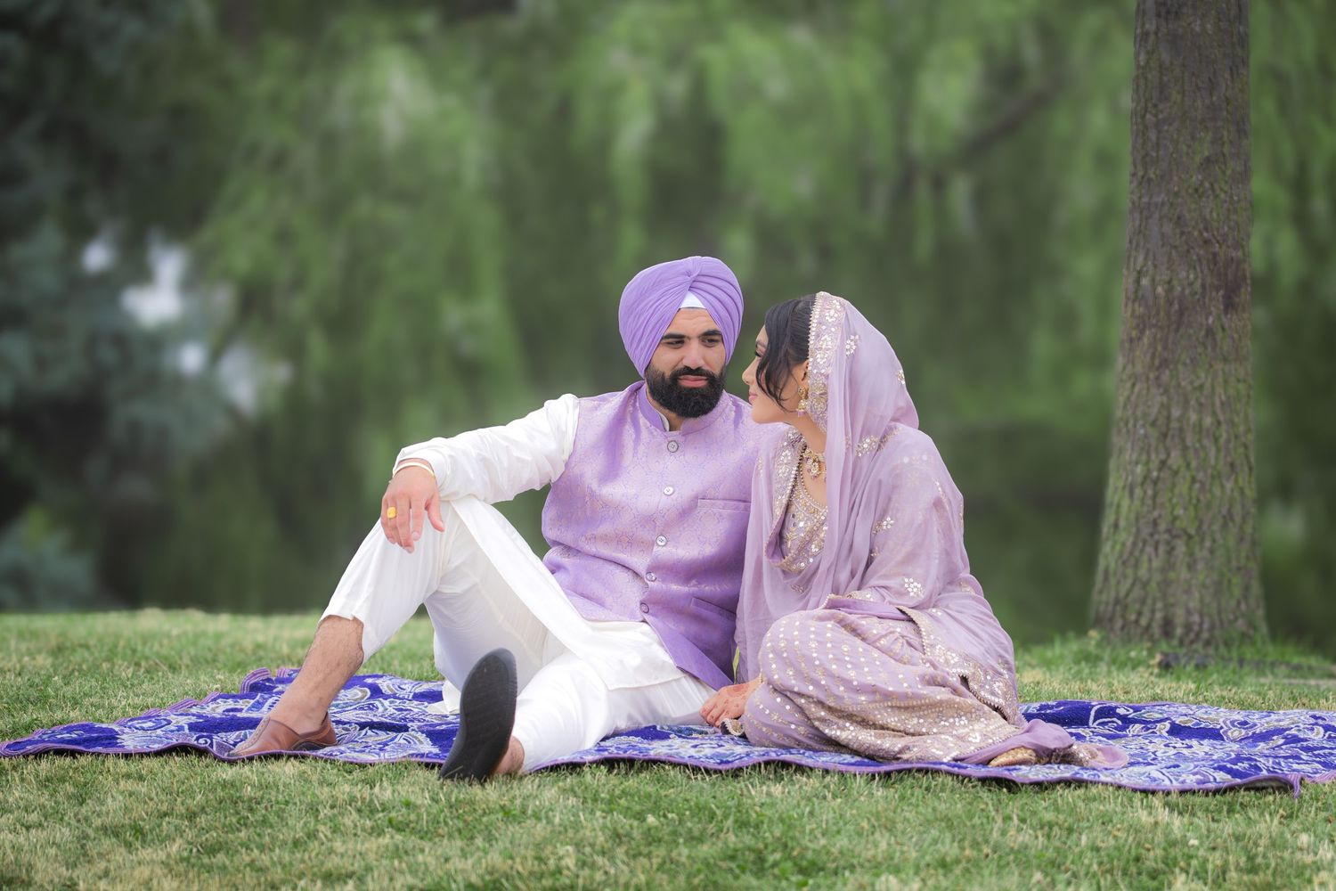 A couple relaxes on a blue patterned blanket in a lush green park setting wearing coordinated purple and white outfits.