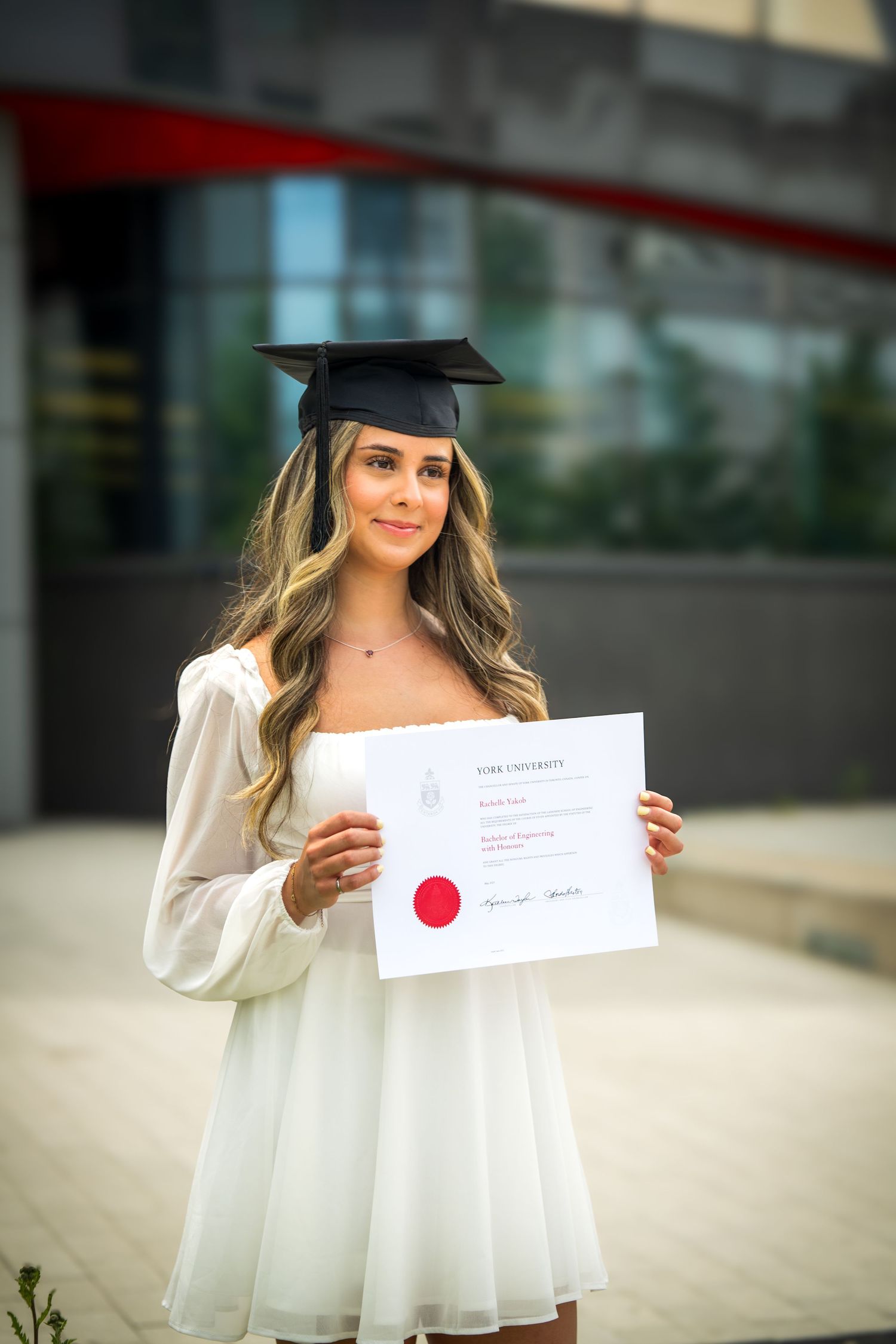 Graduate in white dress and mortarboard displays academic diploma outdoors.