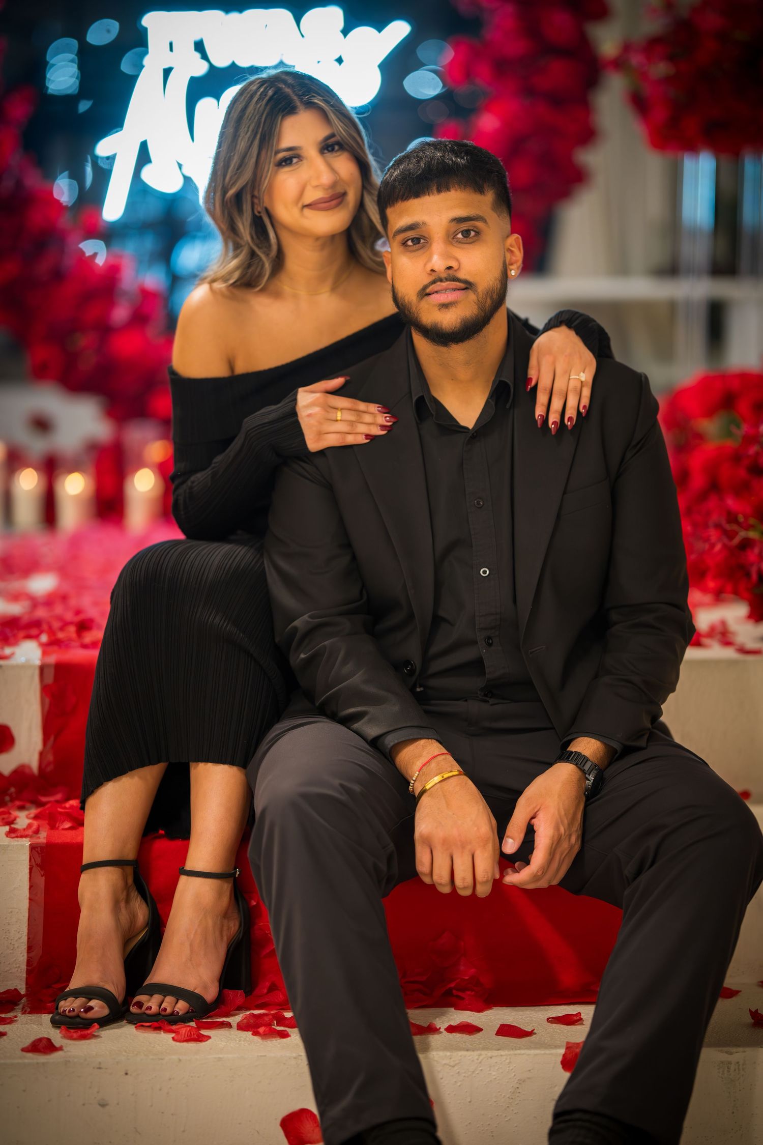 Couple poses in elegant black attire against romantic red rose wall setting.