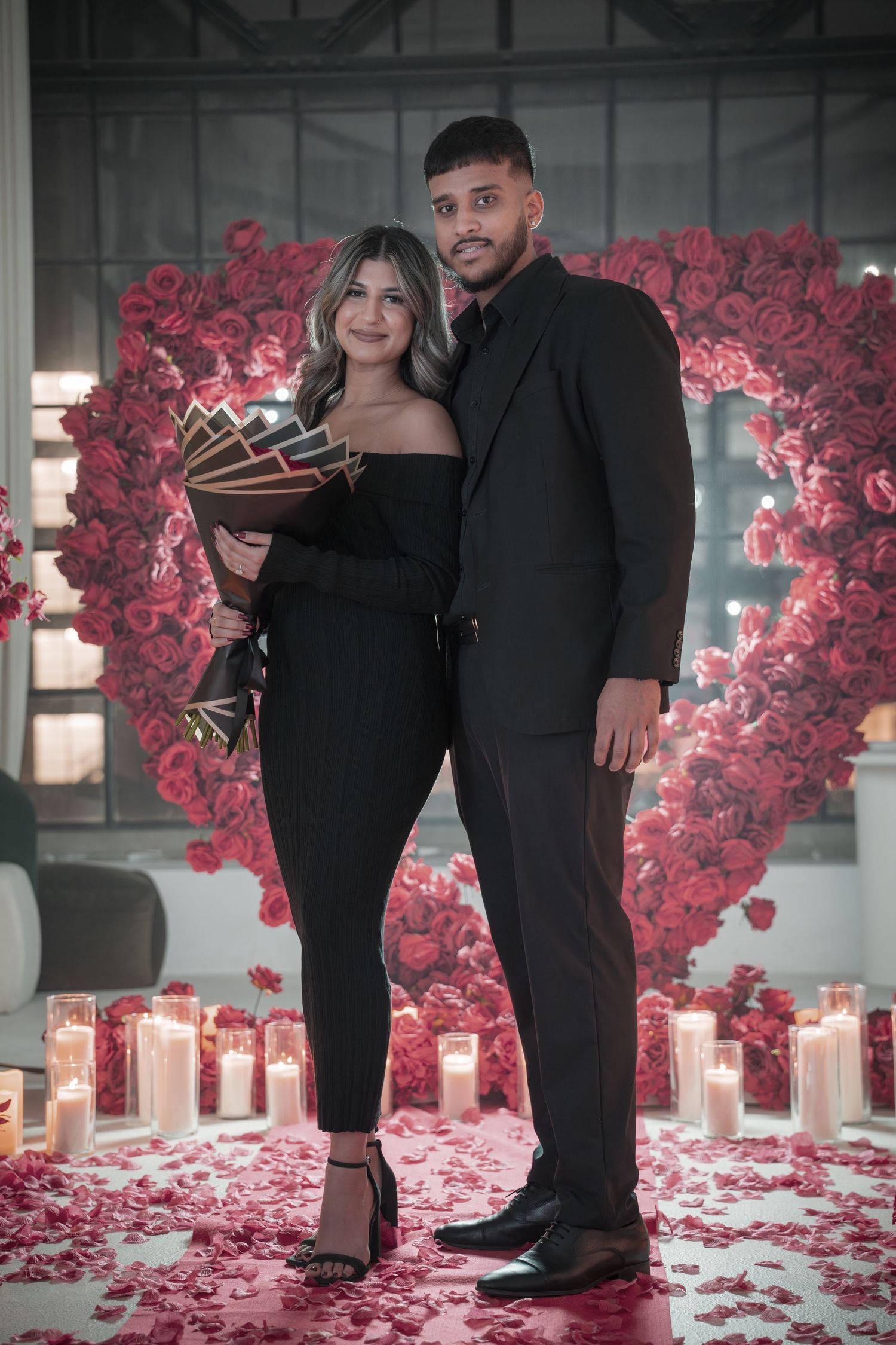 Couple in black attire standing against backdrop of red roses and candles on the floor.