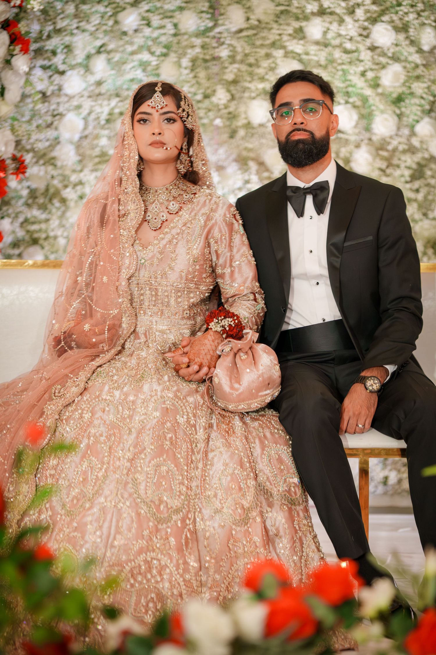 Formal wedding portrait of couple in traditional South Asian attire against floral backdrop.
