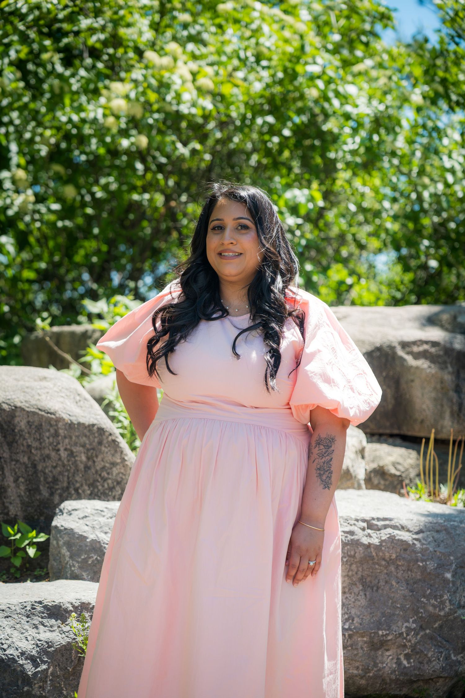 Woman in pink puff-sleeve maxi dress posing outdoors near stone rocks with green trees in background.