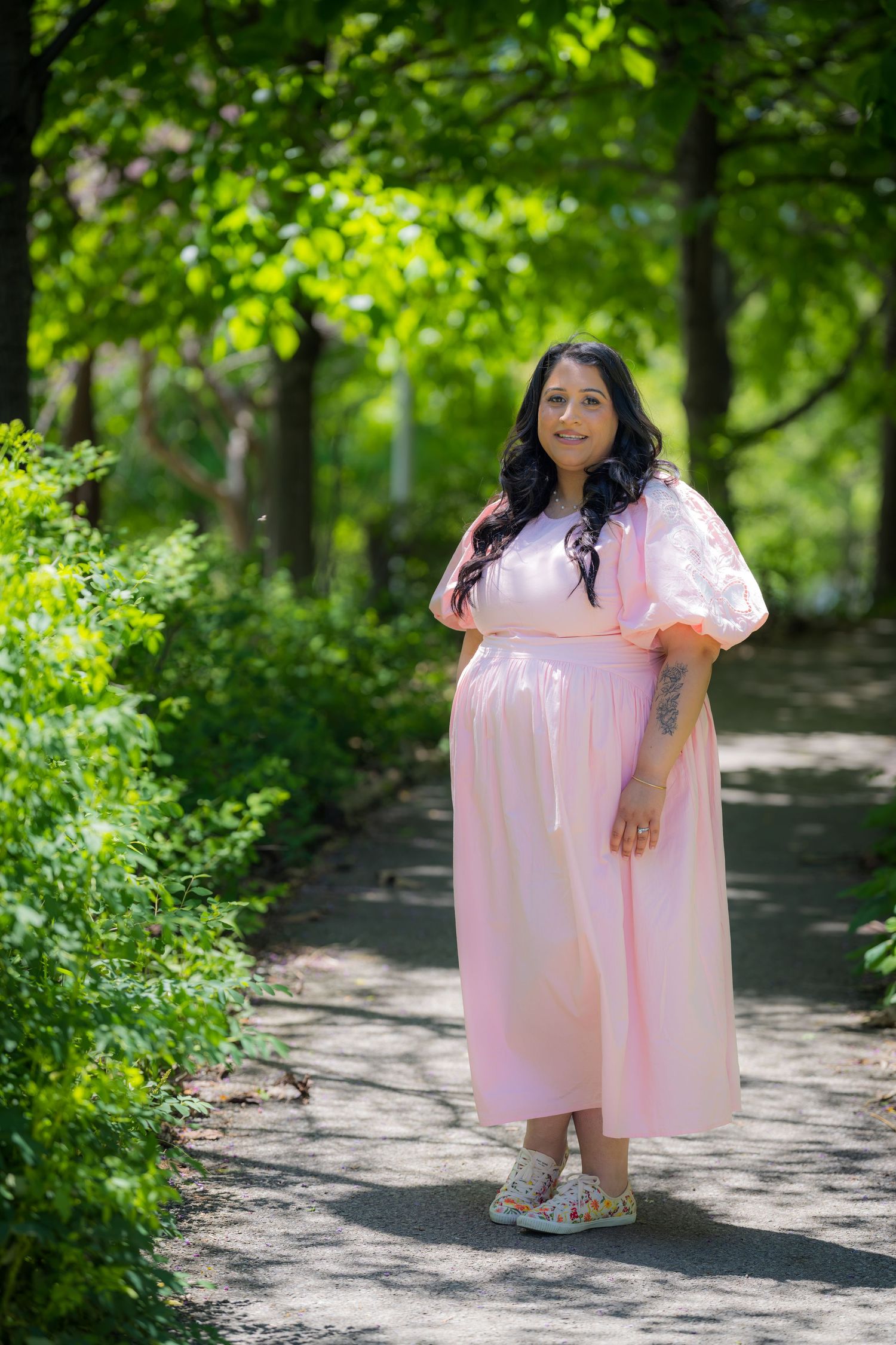 A pregnant woman in a pink maxi dress poses on a tree-lined garden path in sunlight.