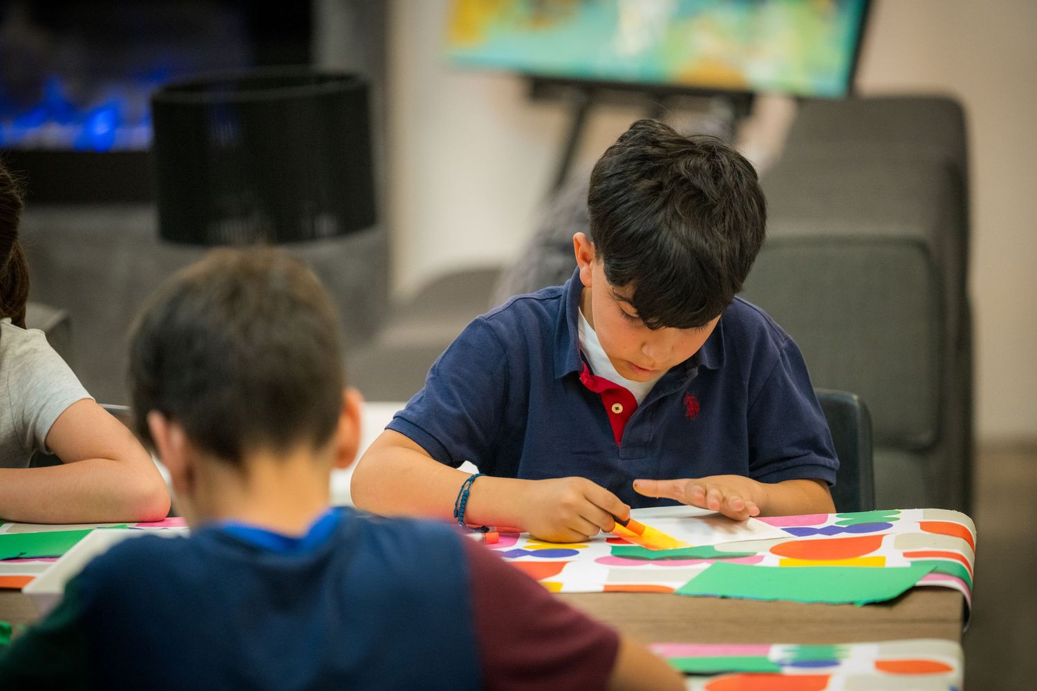 A boy in a blue polo shirt carefully works on a colorful paper collage art project at a table.