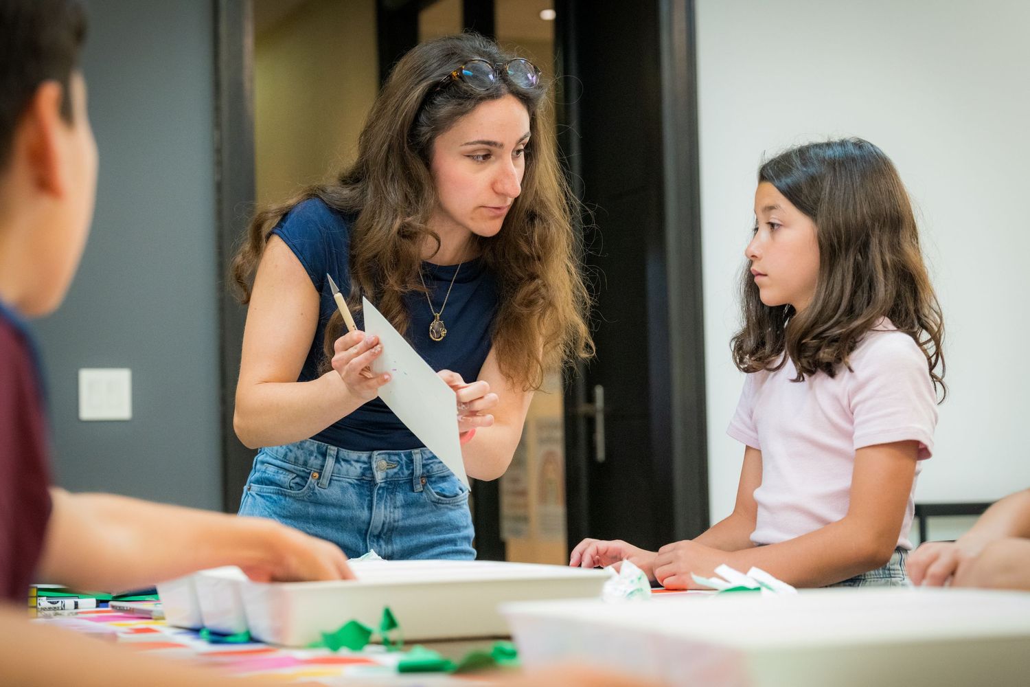 A female art instructor shows a paper to a young girl during a creative art workshop.