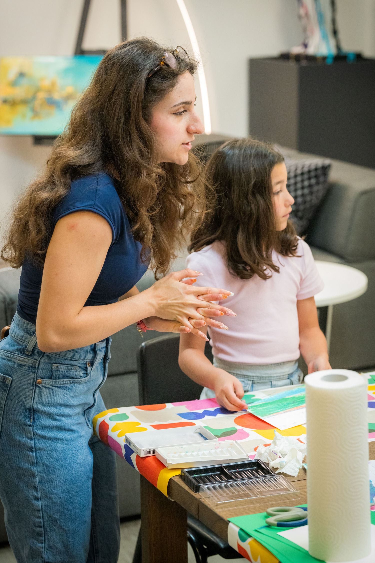 Art teacher explains project details to young girl at a colorful craft-covered table in studio.
