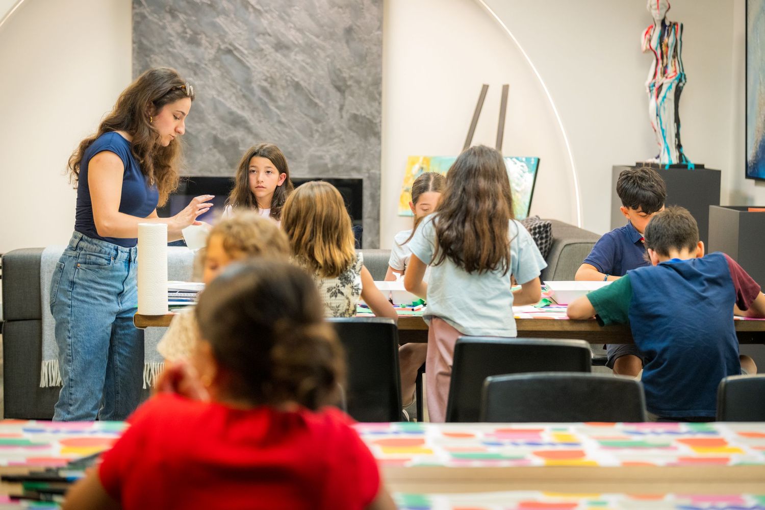 Teacher guides children in an art class as kids paint and create at a colorful table in a bright studio.