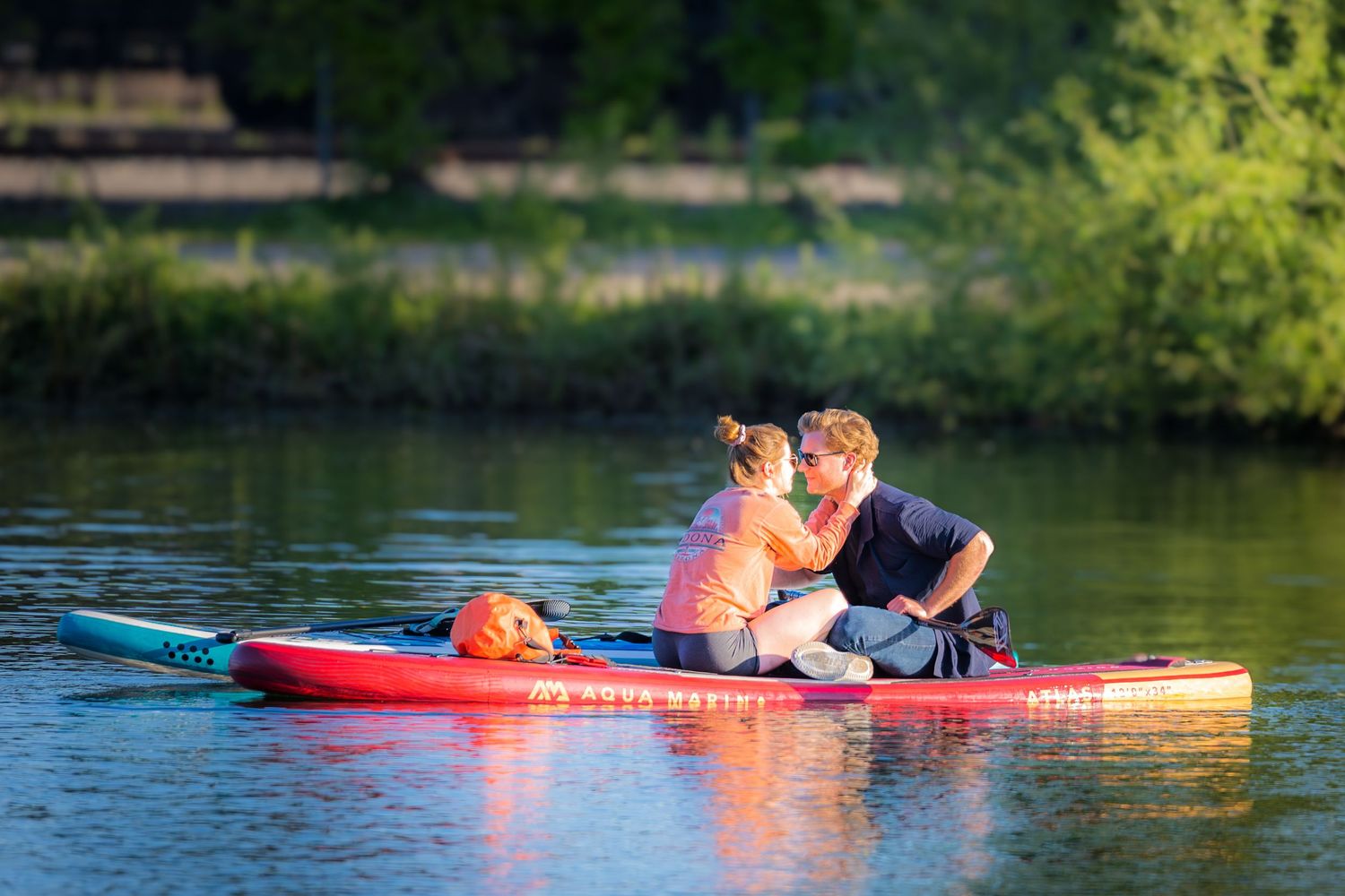 Couple sharing a kiss while sitting together on a red paddleboard on a calm river at sunset.