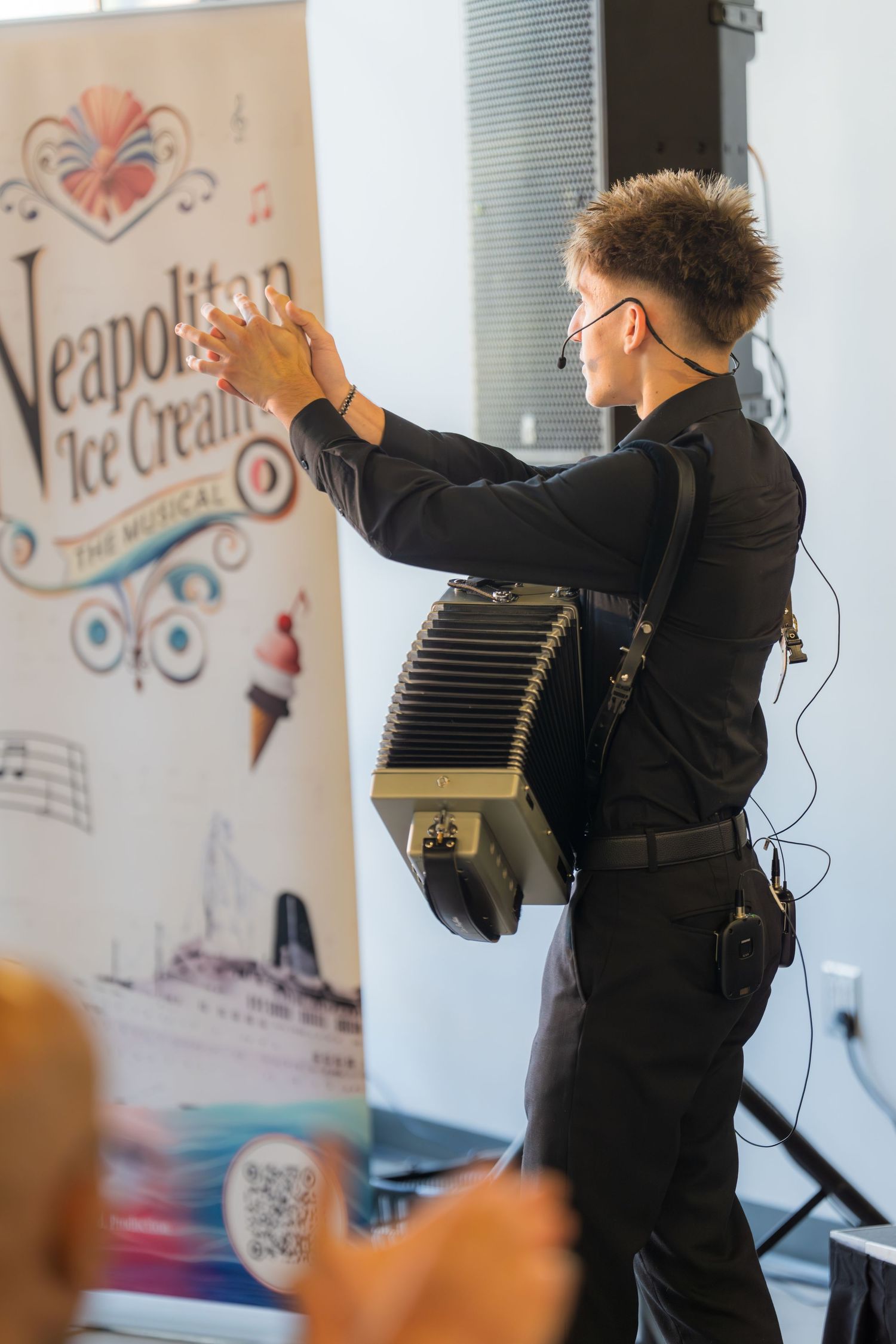 Accordion player in black poses beside Neapolitan Ice Cream The Musical promotional banner.