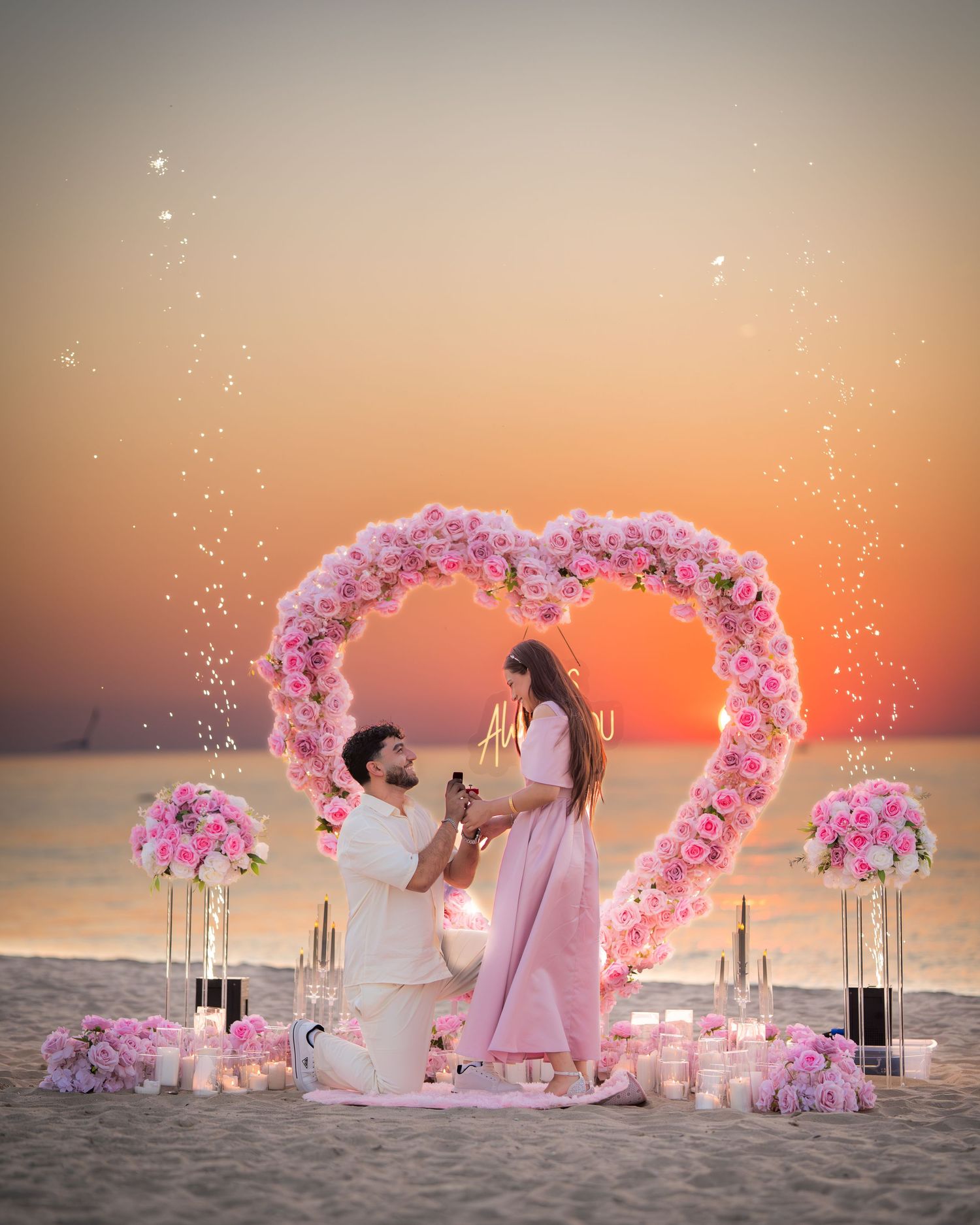 A romantic beach proposal setup with a heart-shaped pink floral arch and candles glowing against a vibrant sunset.