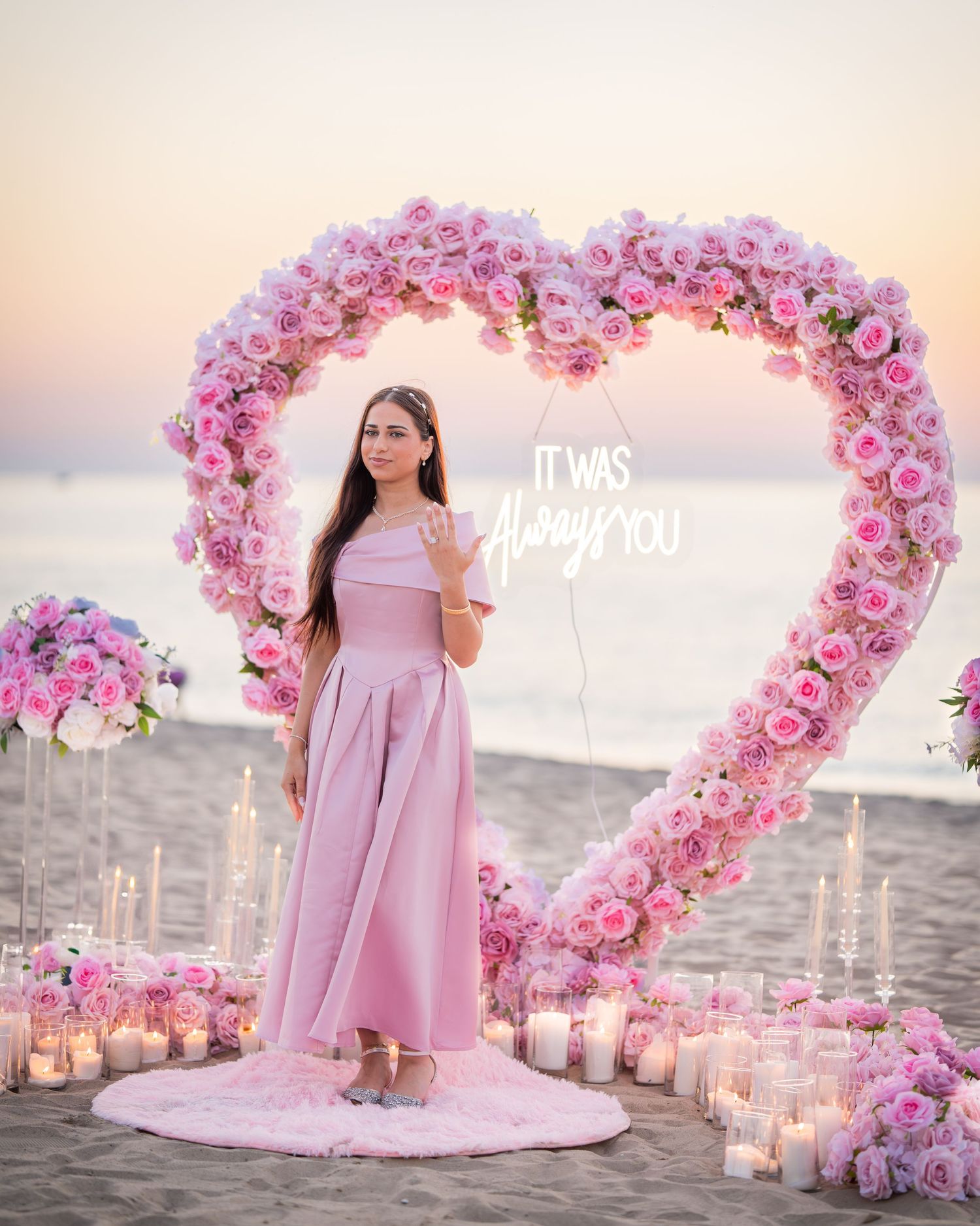 Dreamy beach proposal scene with pink floral decorations and candles lining the sand at sunset.
