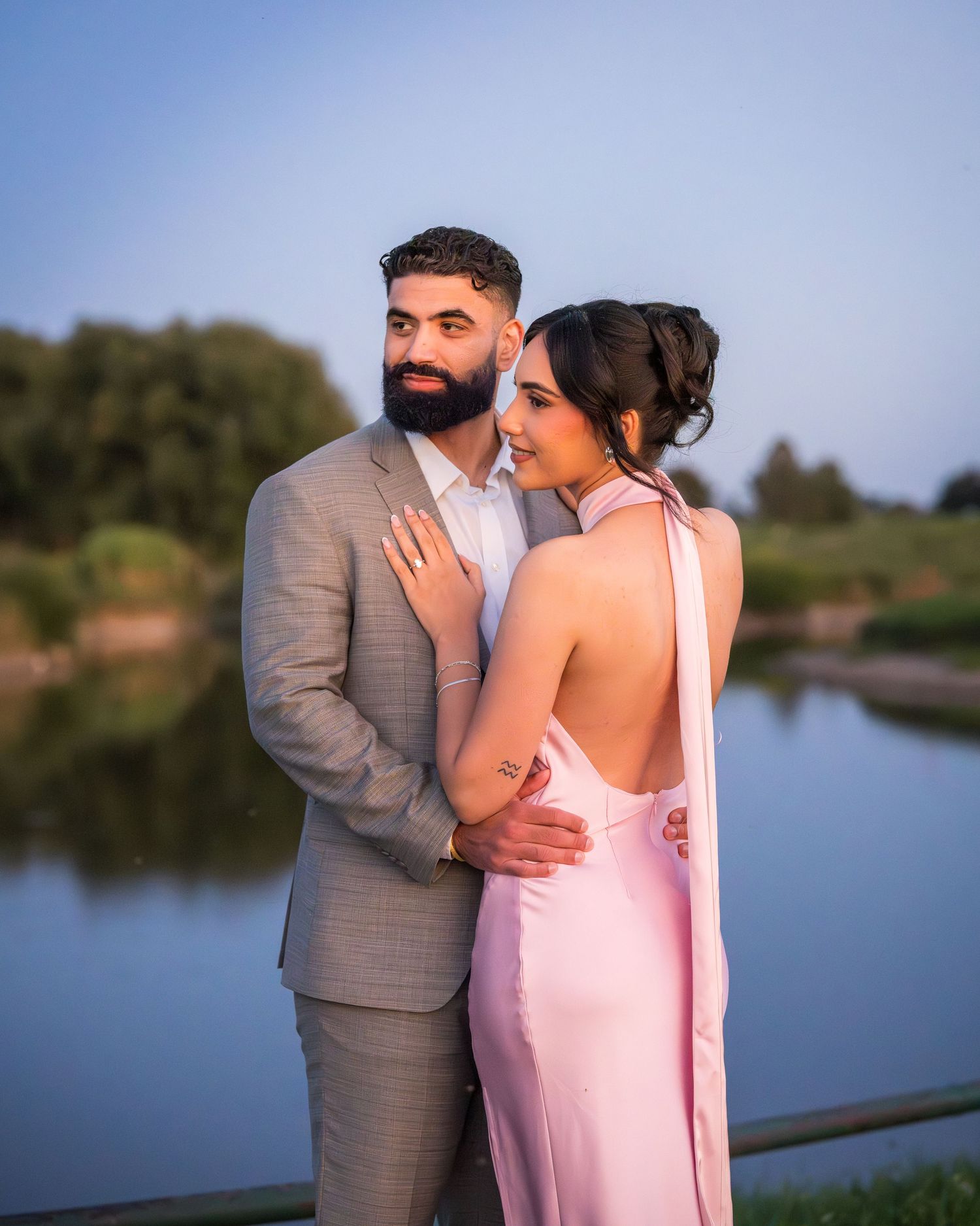 A romantic sunset photoshoot by a lake with a couple in pink dress and grey suit embracing.