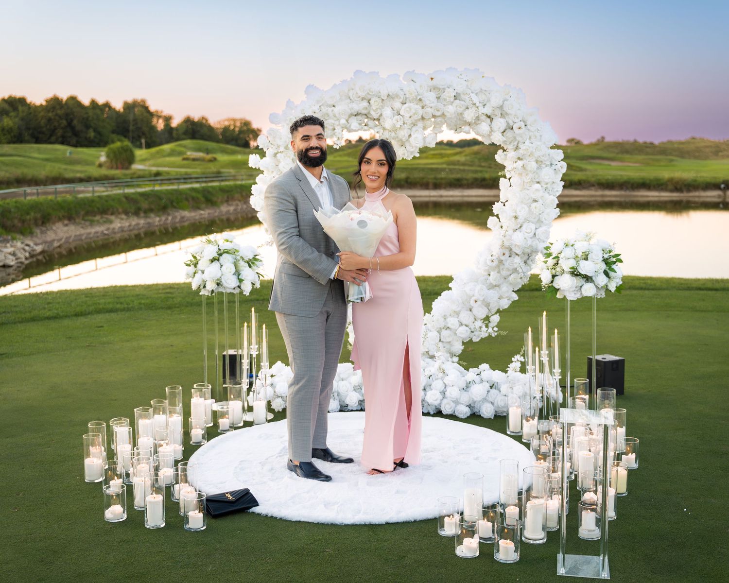 Elegant outdoor proposal scene with white floral arch and glowing candles on manicured lawn.