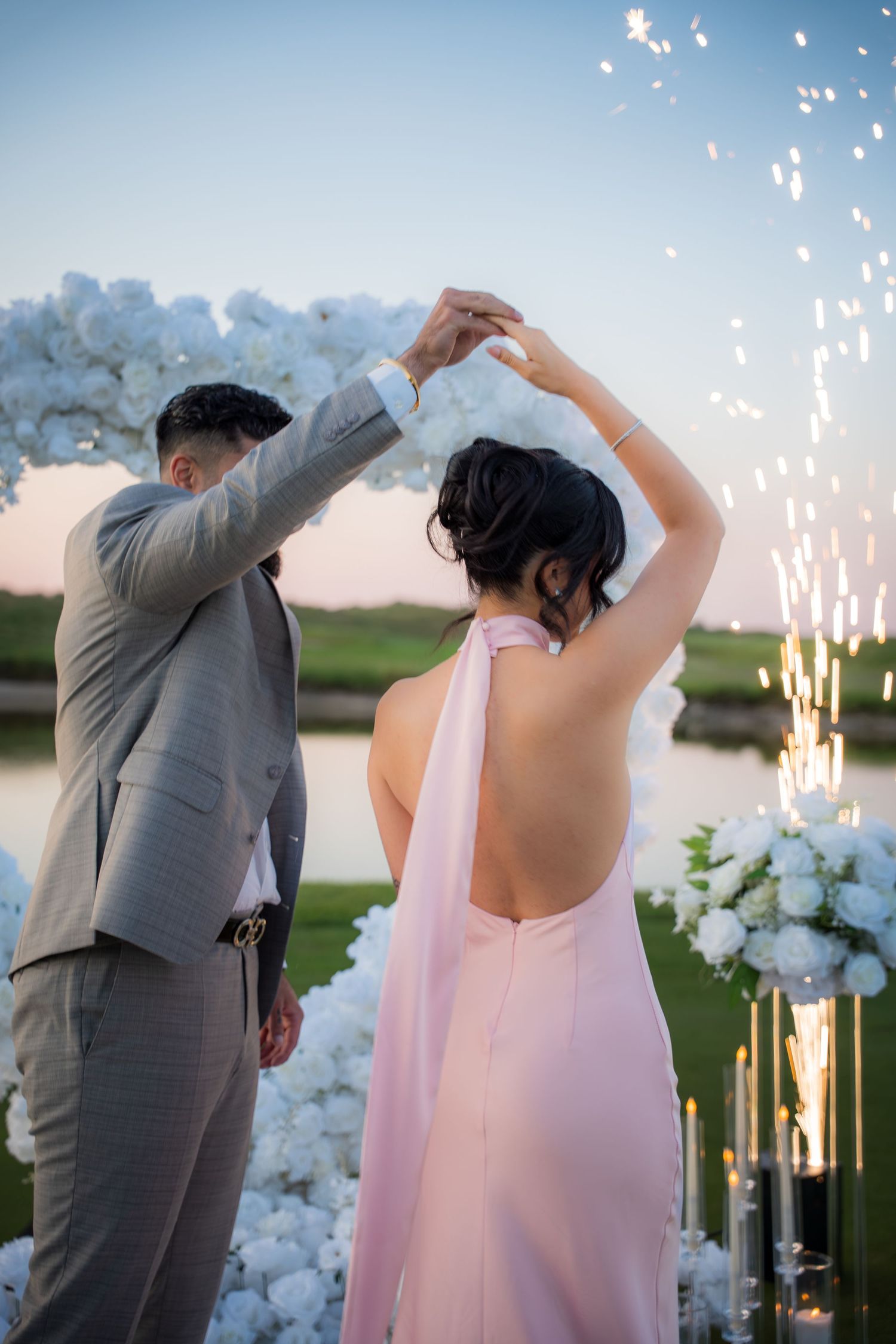 Dancing couple silhouetted against sparklers and floral arch during sunset wedding celebration.