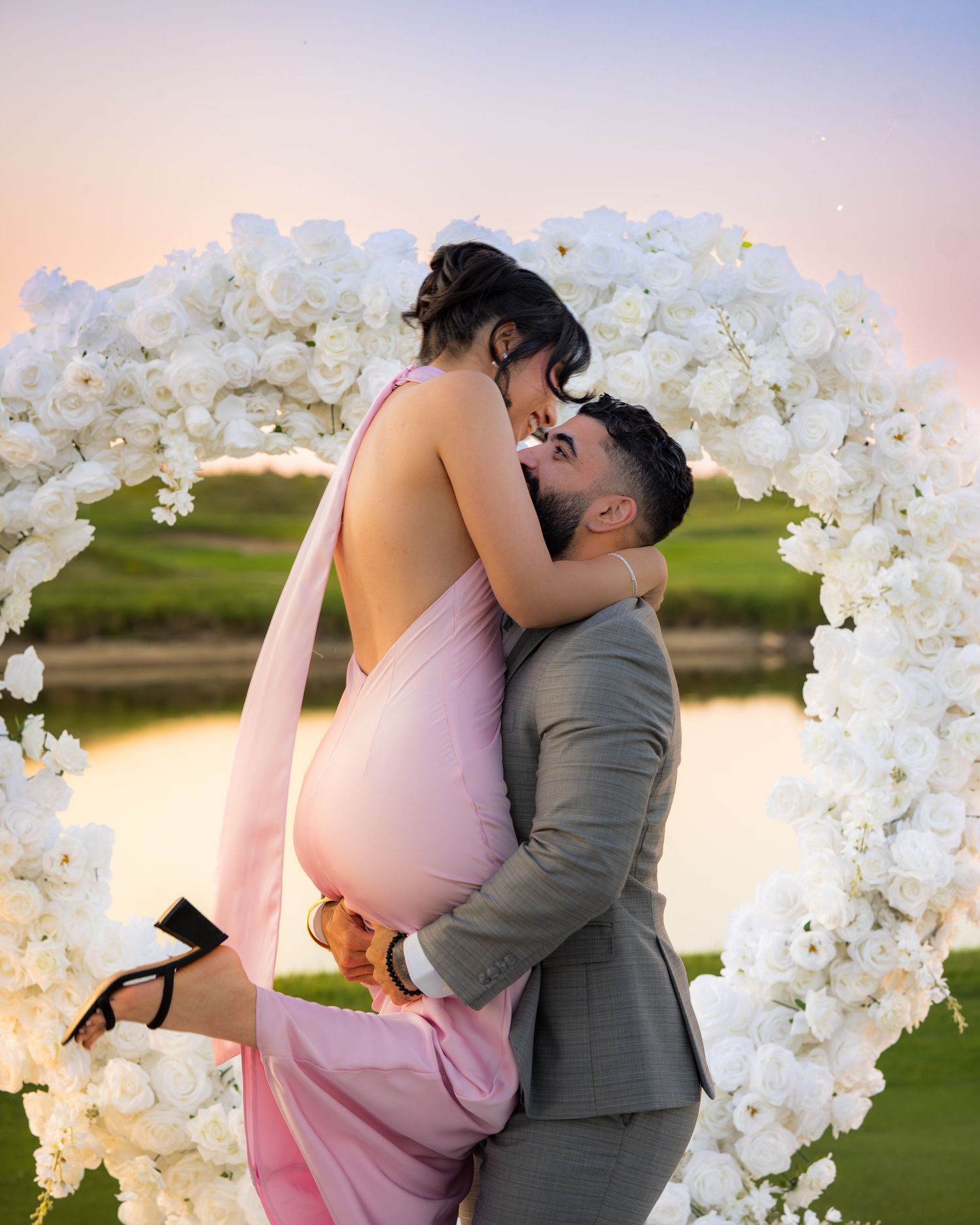 A romantic kiss between a couple wearing pink and grey attire beneath a floral arch at dusk.