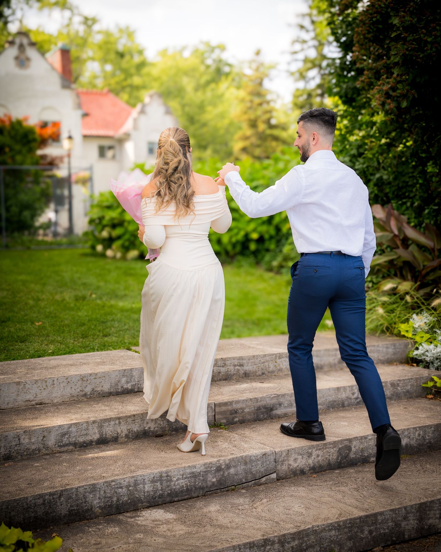 A couple in wedding attire playfully walks along a concrete wall in a garden setting.