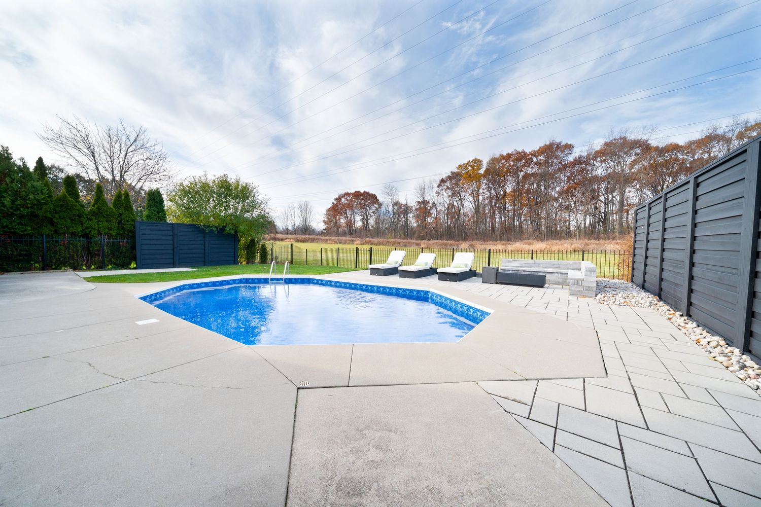 Modern backyard swimming pool with concrete patio deck and privacy fencing under bright blue skies.