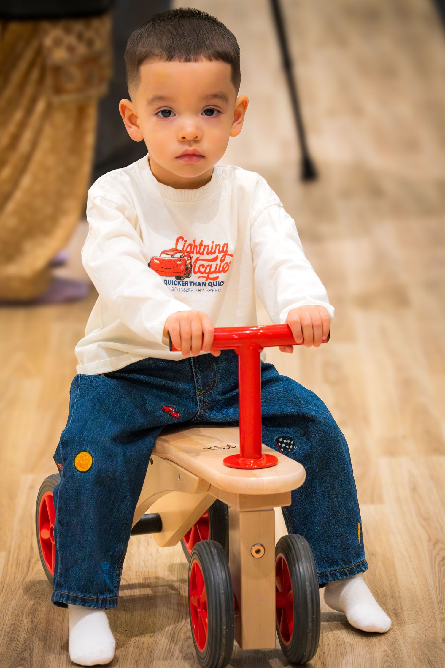 Toddler boy in Lightning McQueen shirt riding a wooden balance bike on hardwood floor.