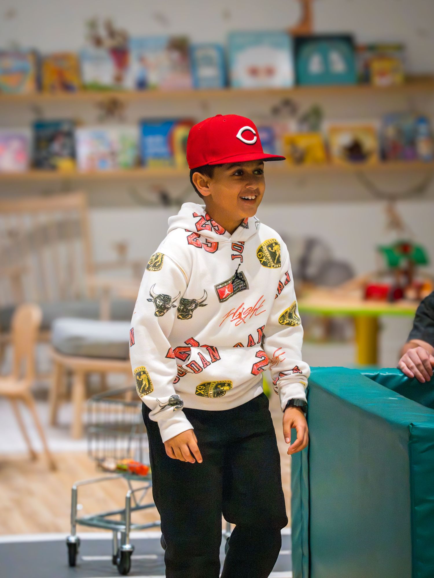 Young boy wearing a red Cincinnati Reds cap and graphic hoodie smiling in a colorful children's play area.