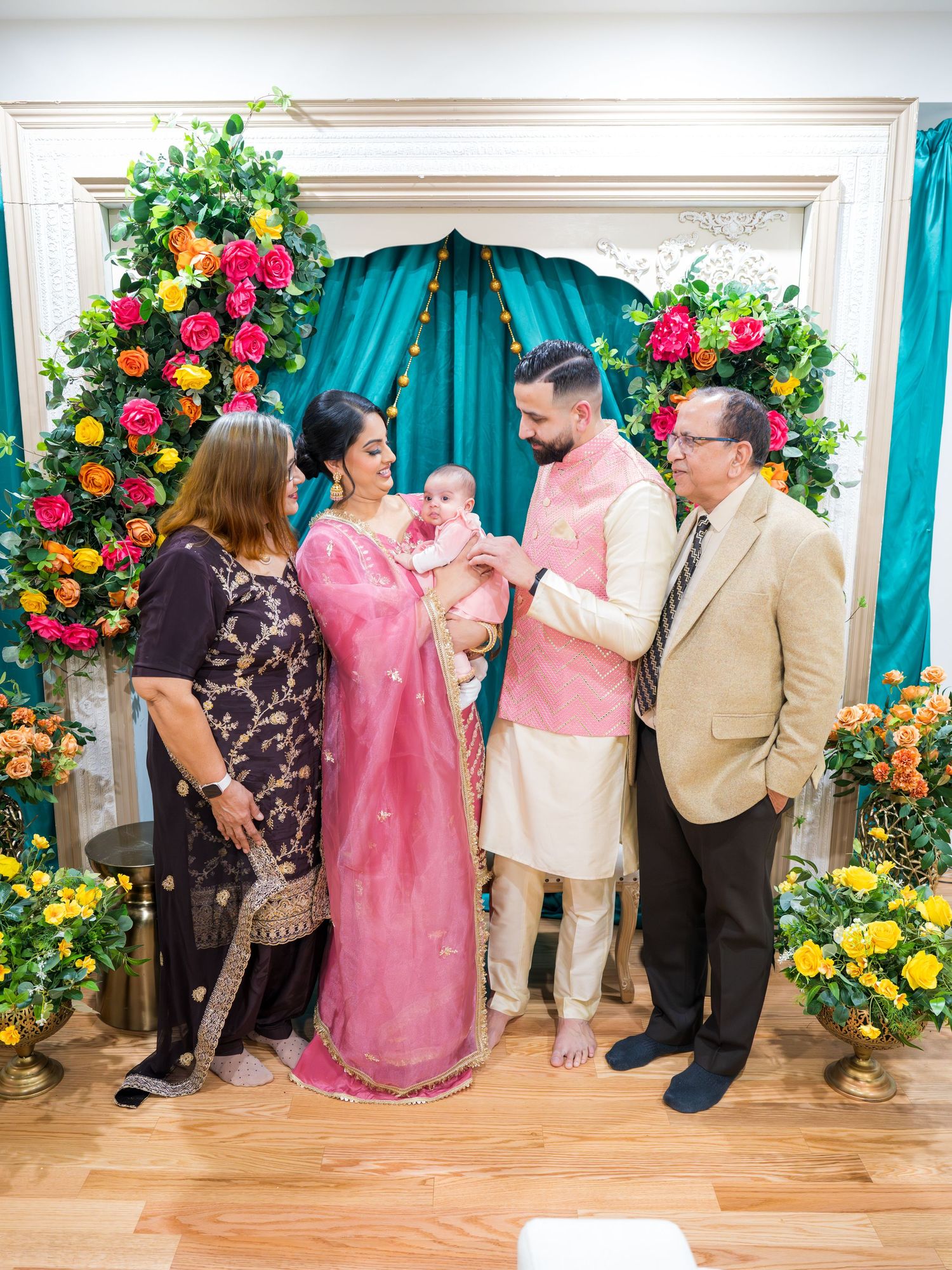 Family of four with baby celebrating South Asian ceremony with floral arch and teal drape backdrop.