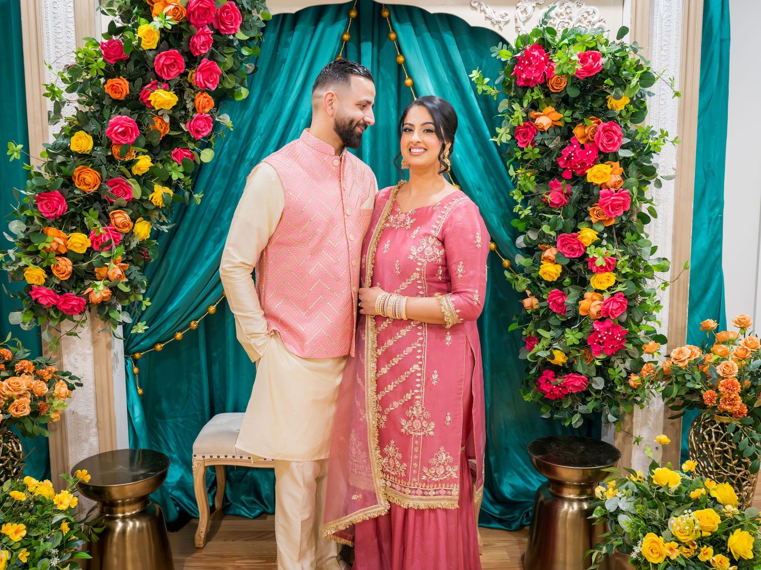 A couple in pink Indian traditional attire poses together in front of a colorful floral arch decoration.