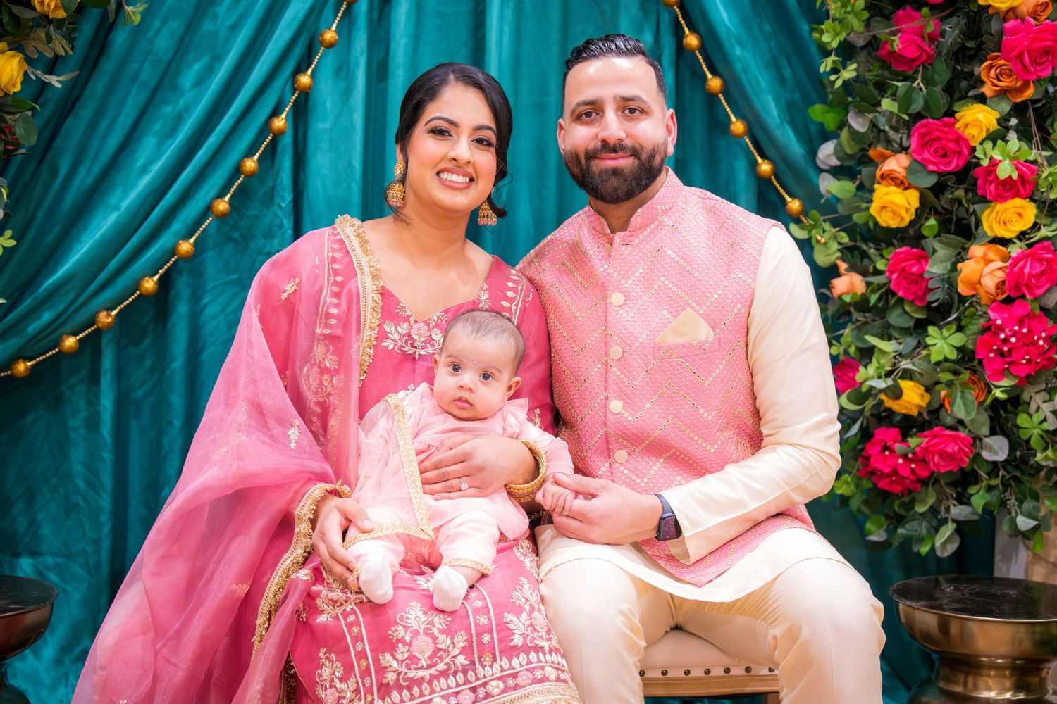 Smiling Indian couple in matching pink ethnic wear sits with infant at festive cultural ceremony.
