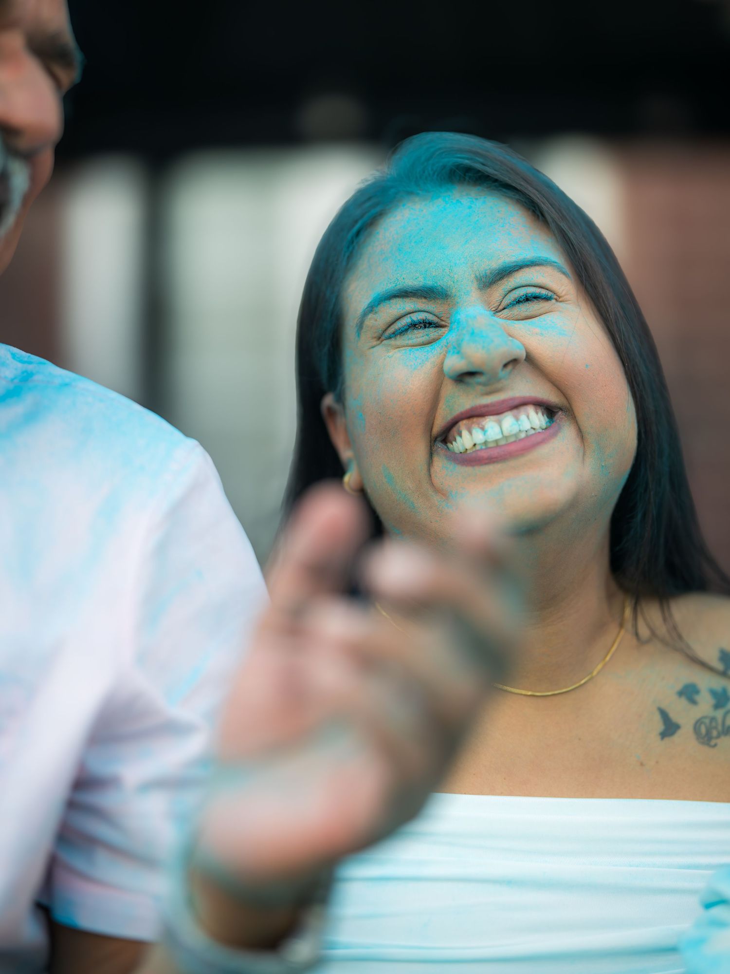 A woman laughs joyfully with blue gender reveal powder on her face at an outdoor celebration.