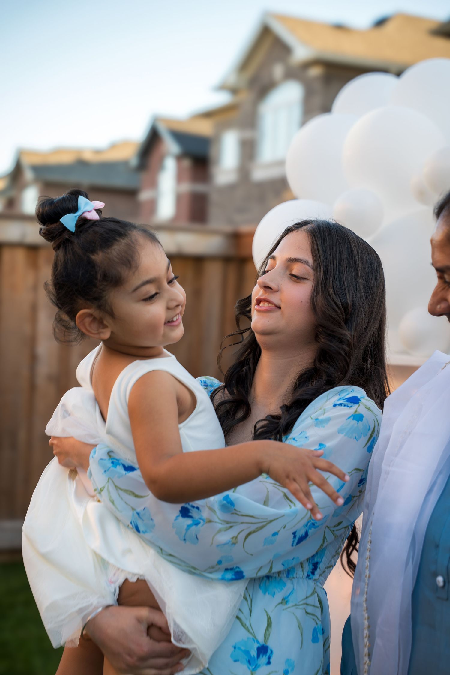 A young woman in a floral dress smiles warmly while holding a toddler girl near white balloons outdoors.