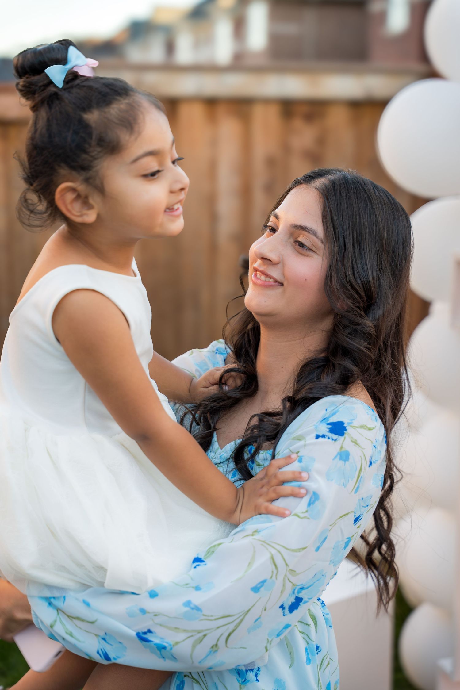 Smiling woman in blue floral dress holds young girl in white dress at outdoor celebration with balloons.