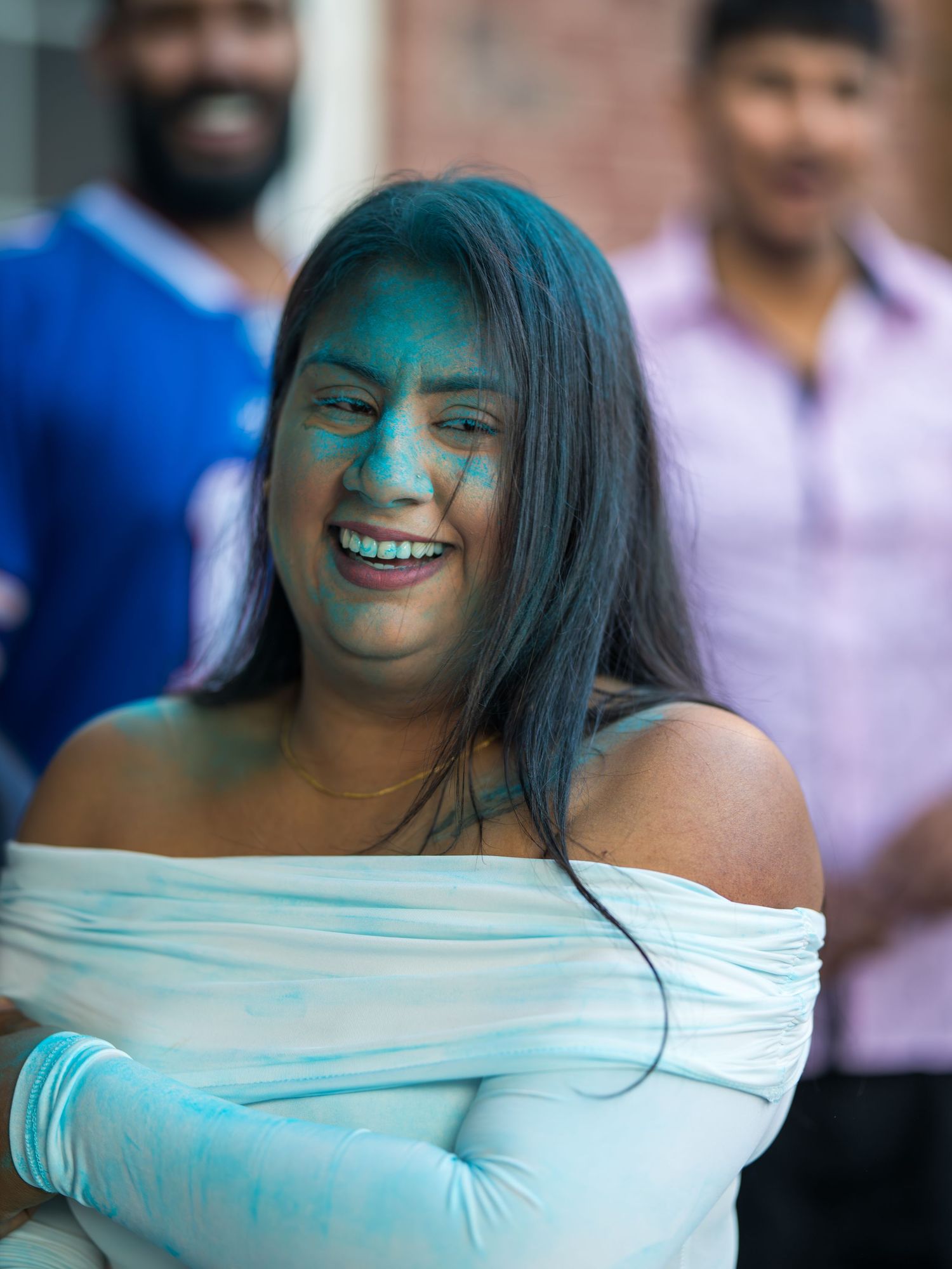 A joyful woman with blue Holi powder on her face wearing a white off-shoulder top at an outdoor festival.
