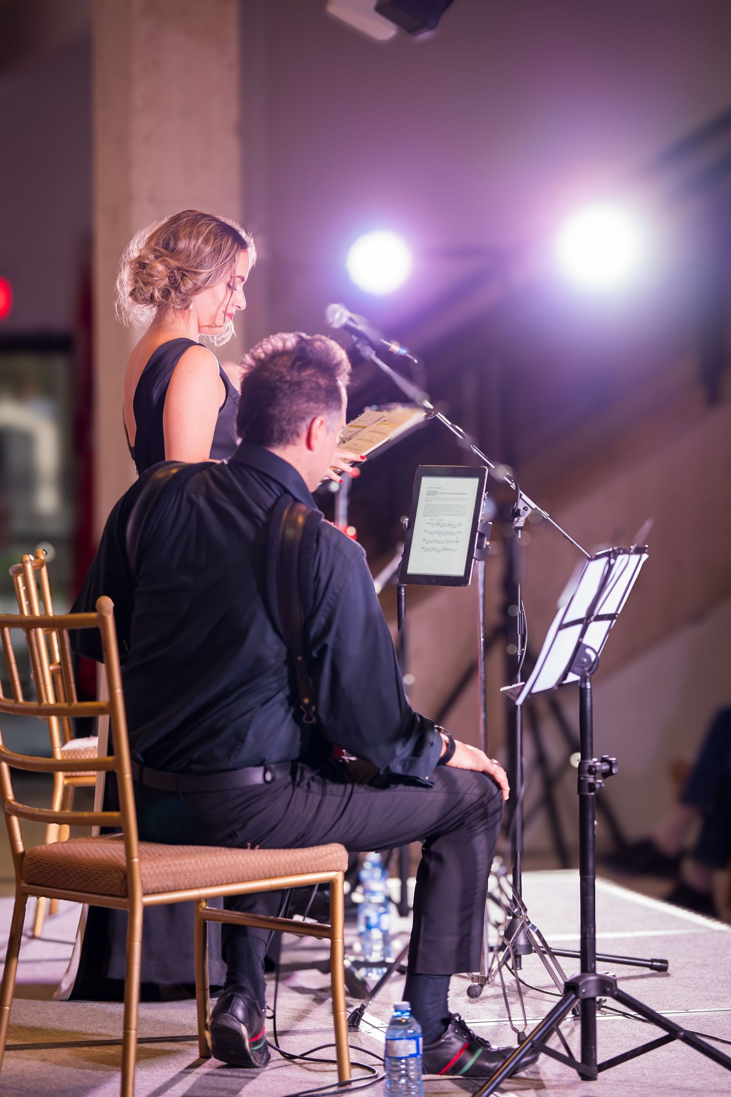 Two musicians in black attire rehearse on stage with microphone stands and music stands under spotlights.
