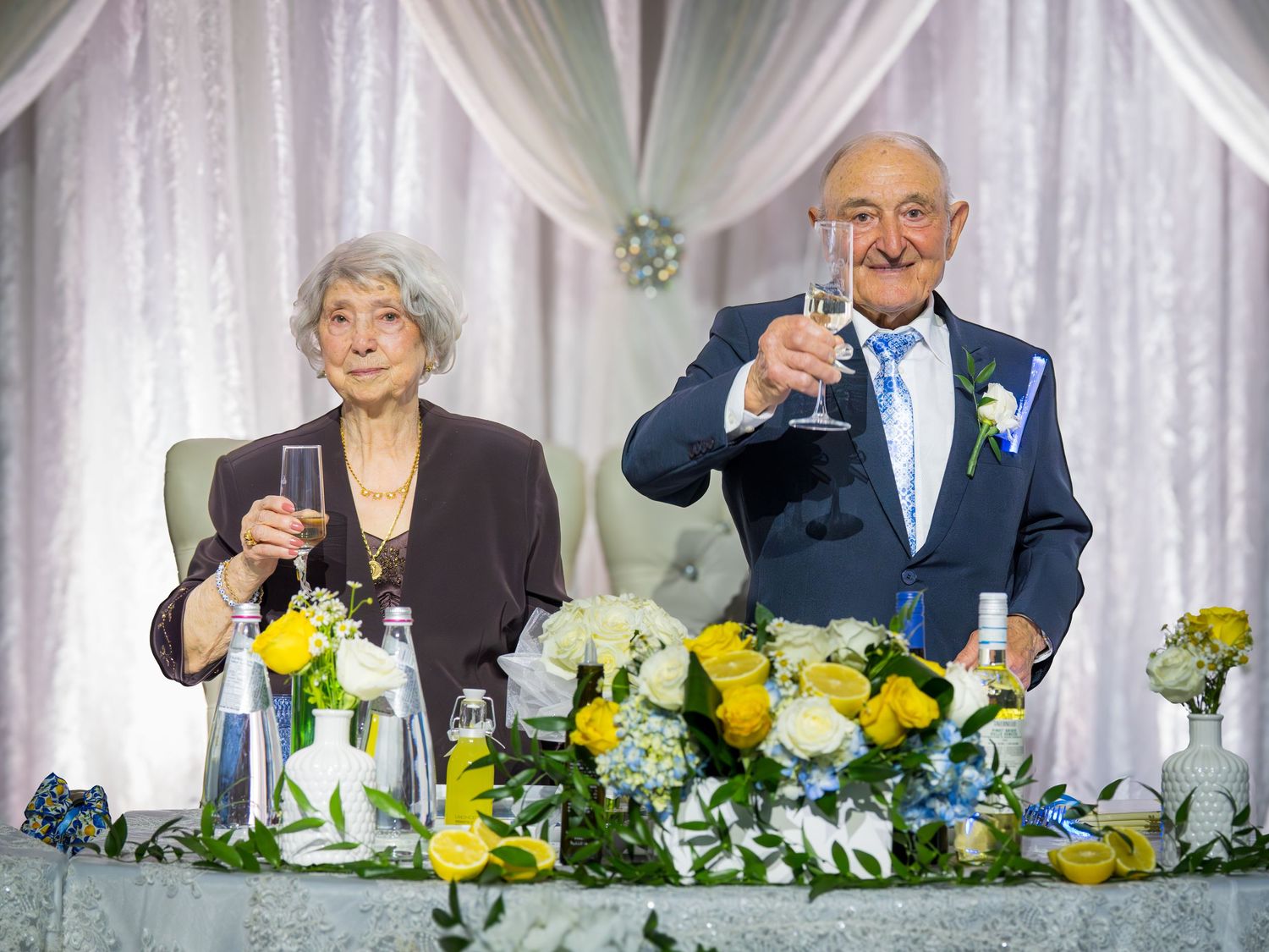 Elderly couple toasting with champagne glasses at a wedding reception with yellow and blue floral arrangements.