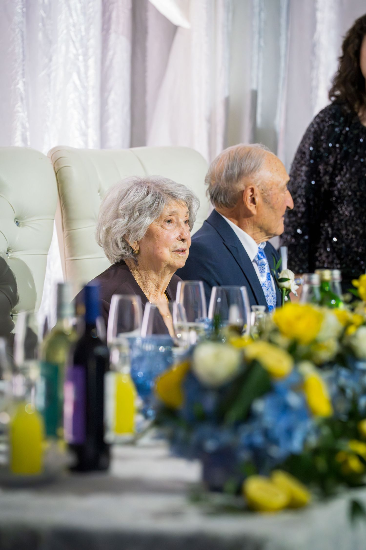 Elderly couple seated at elegantly decorated wedding reception table with yellow and blue floral centerpieces.