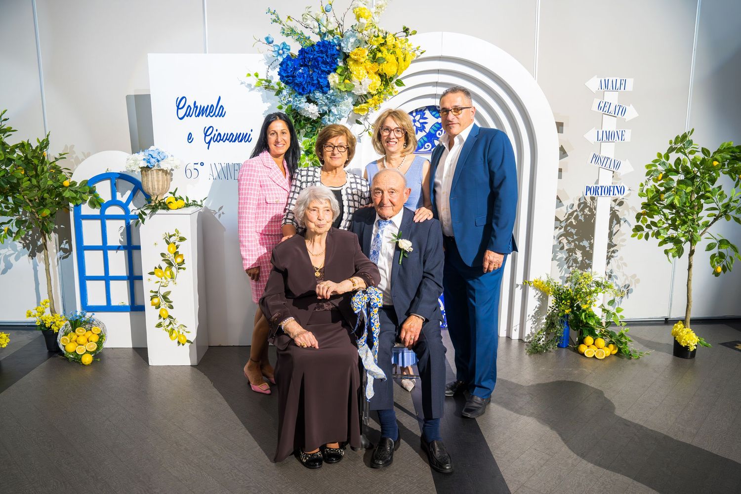 Family group photo in front of a decorative arch reading Carmela and Giovanni at an anniversary celebration.