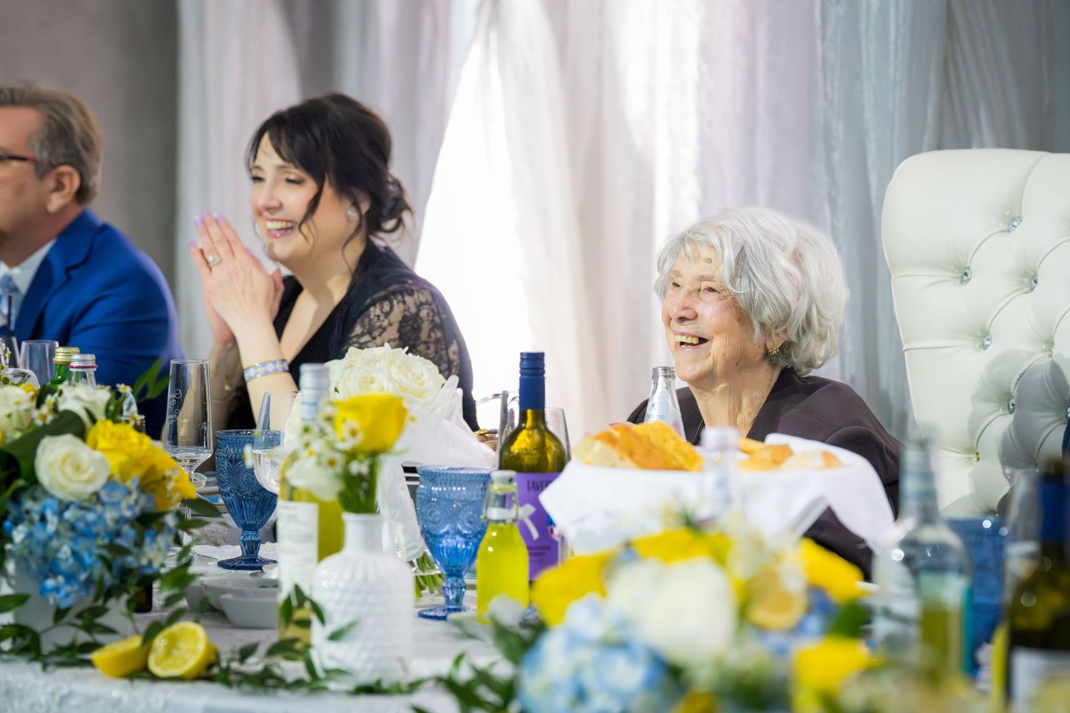 Two women smile and clap at elegantly decorated wedding reception table with floral centerpieces.