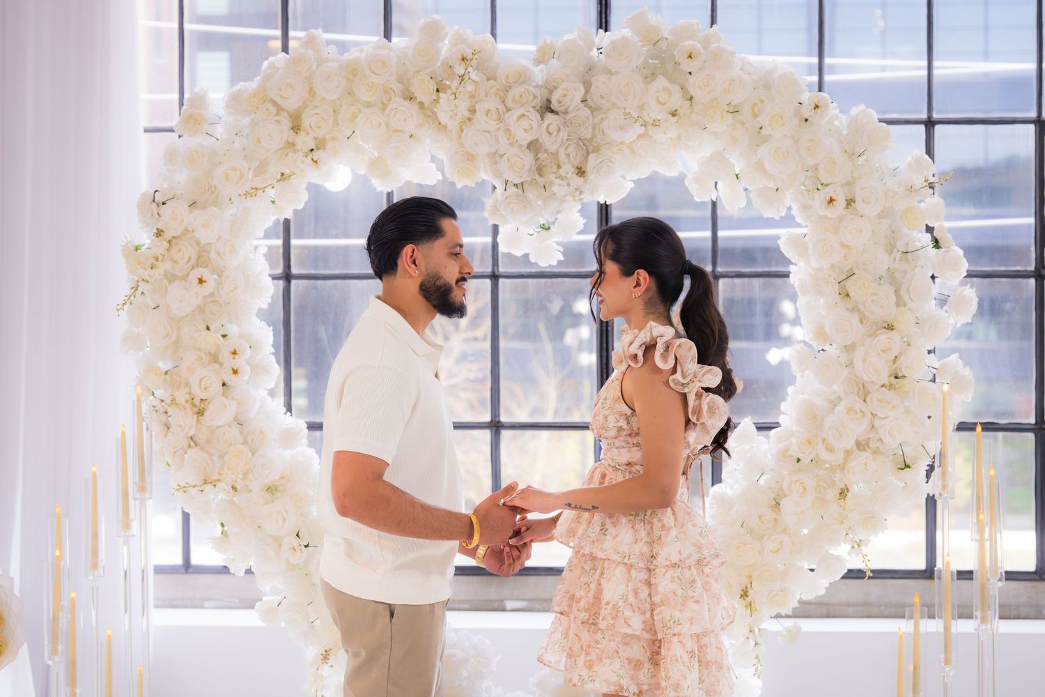 Couple holding hands under a white floral heart arch during a romantic proposal setup.