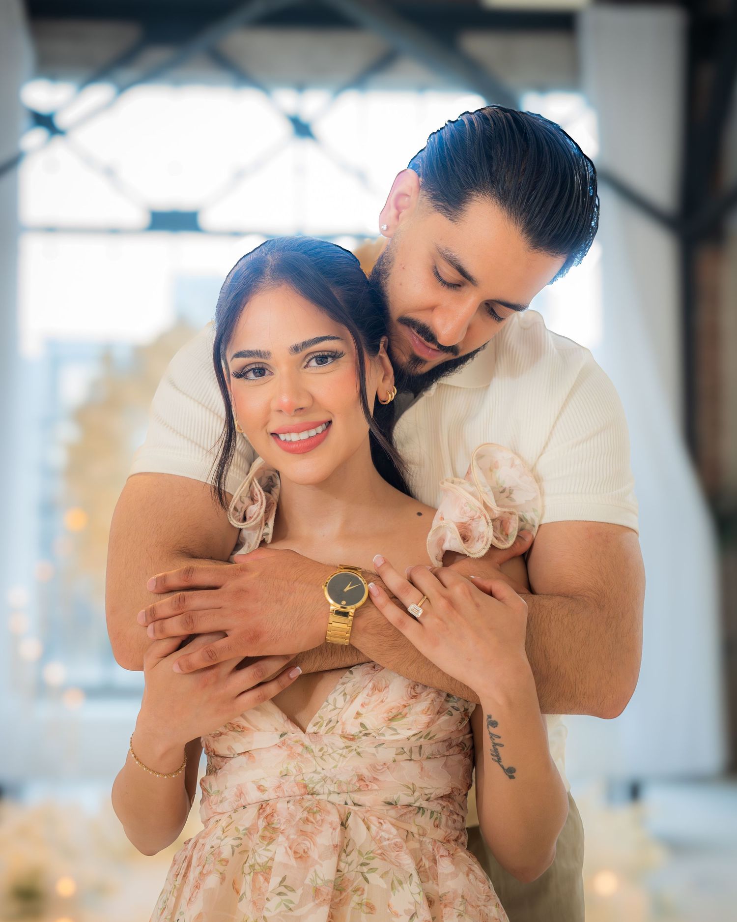 Man embracing woman from behind, both smiling, wearing floral dress and gold watch in romantic setting.