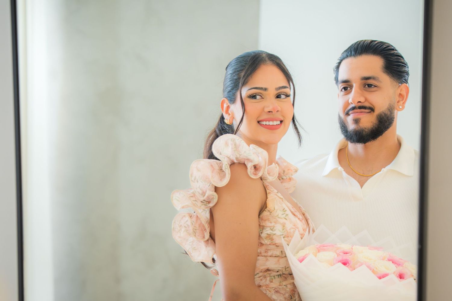 Smiling couple posing together in mirror reflection, woman holding pink bouquet in elegant outfit.