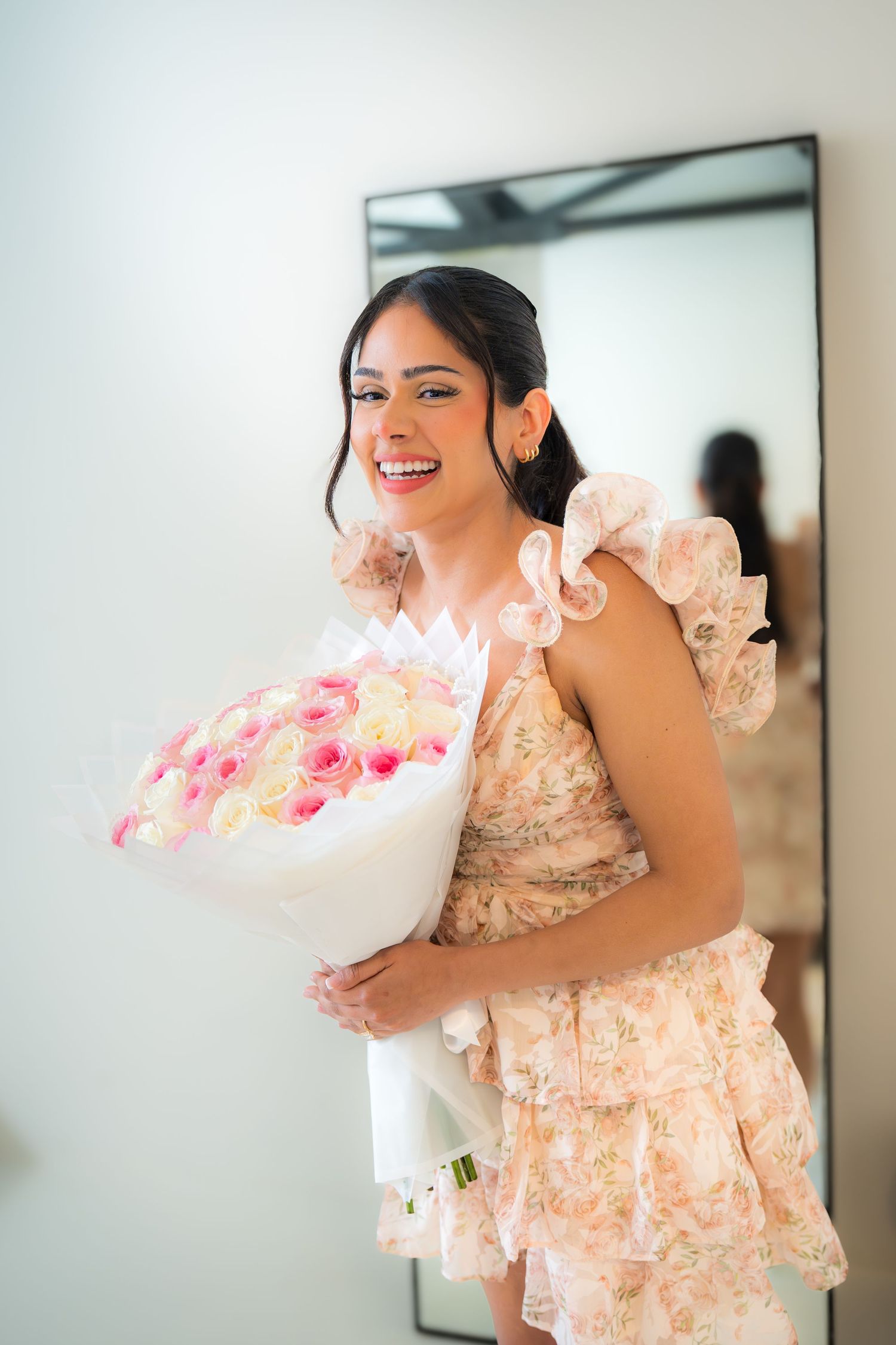 Happy woman in floral ruffle dress holding large bouquet of pink and white roses, smiling joyfully.