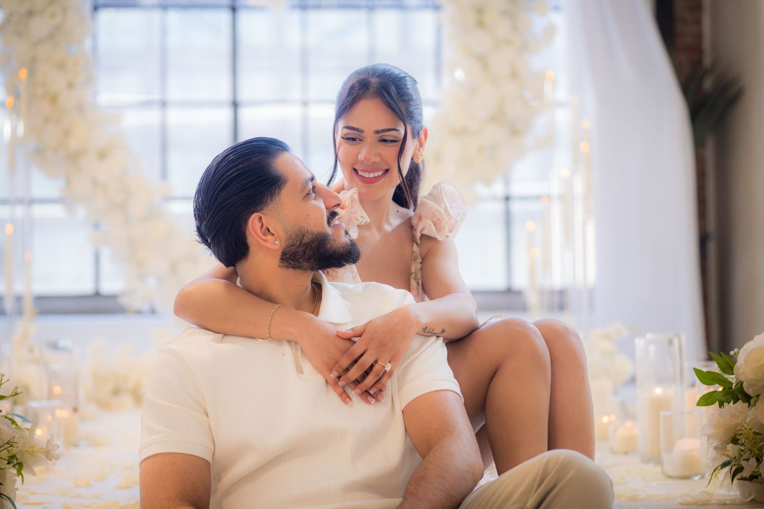 Smiling couple sits together surrounded by candles and white flowers after a romantic proposal.