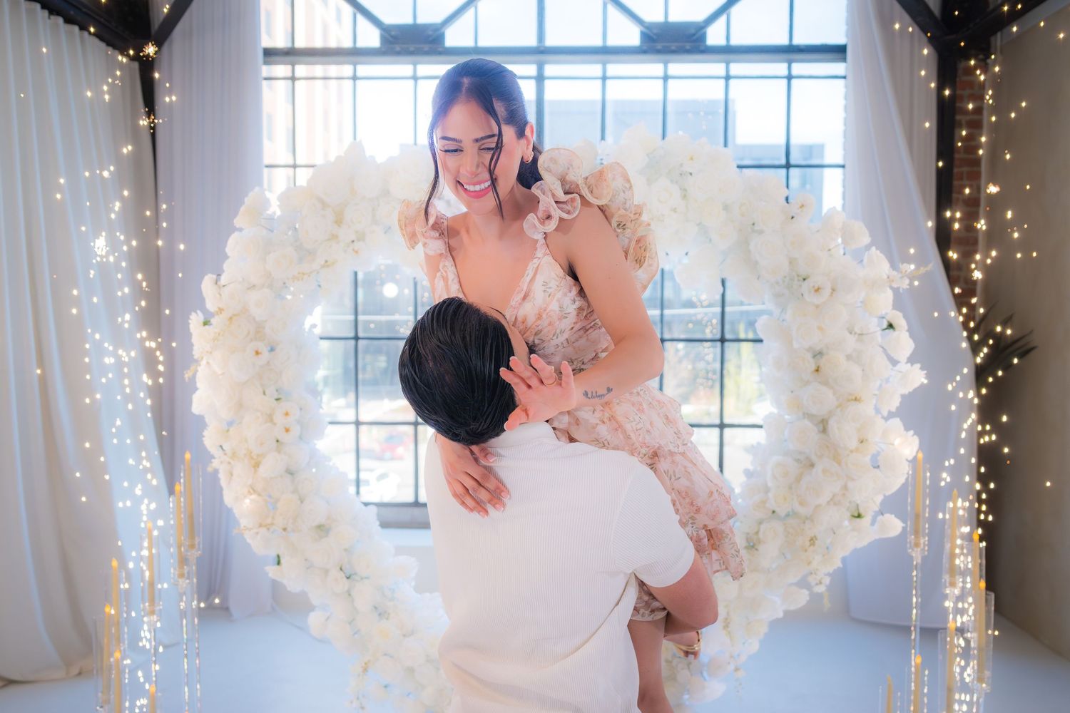 Man lifting his fiancee in front of a stunning white floral heart arch decorated with twinkling fairy lights.